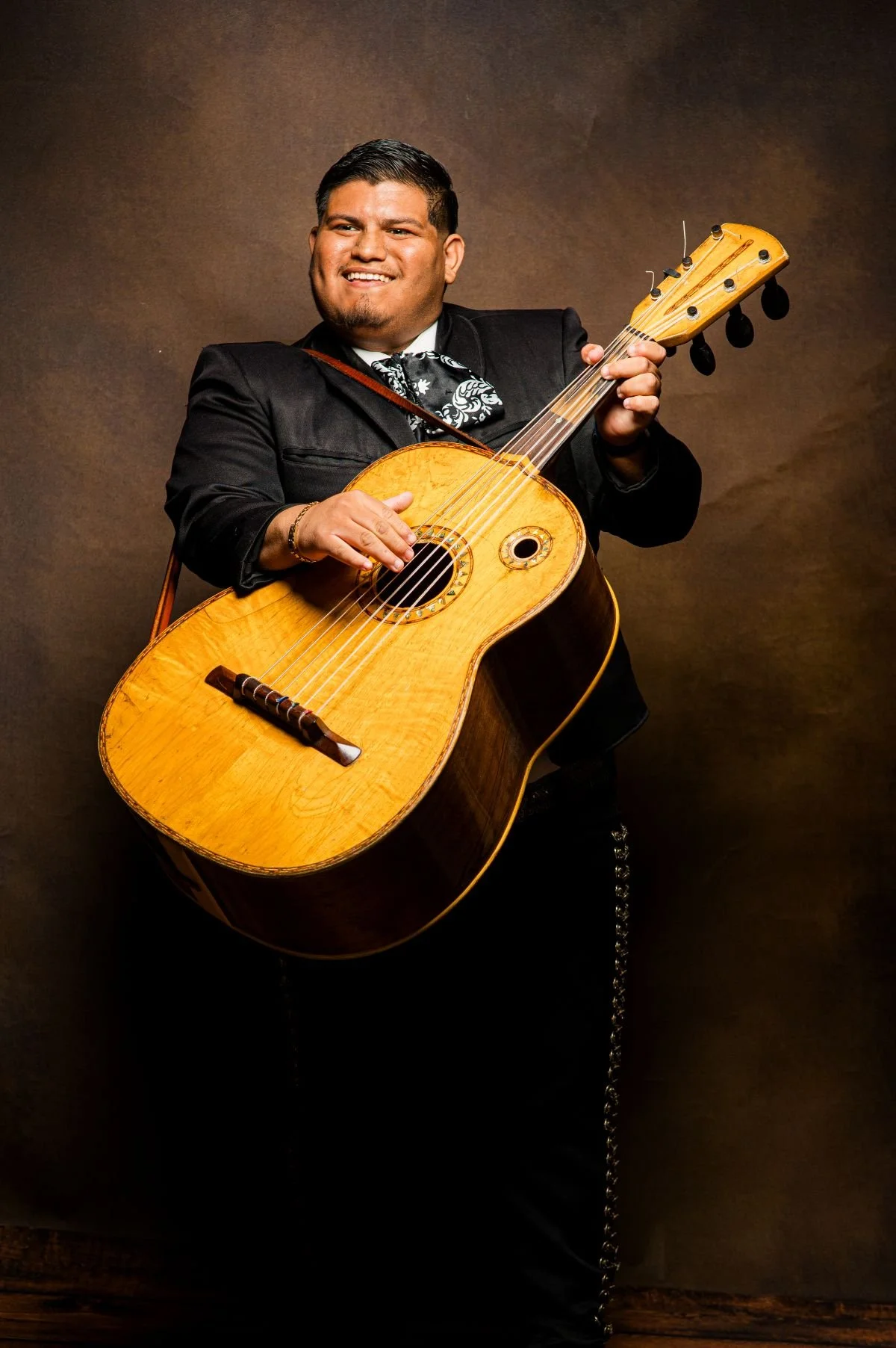A man wearing traditional Mexican attire, holding a wooden acoustic guitar, smiling against a brown background.