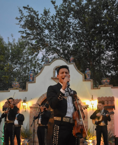 A musician singing and playing violin on an outdoor stage with a group of musicians in the background, during sunset.