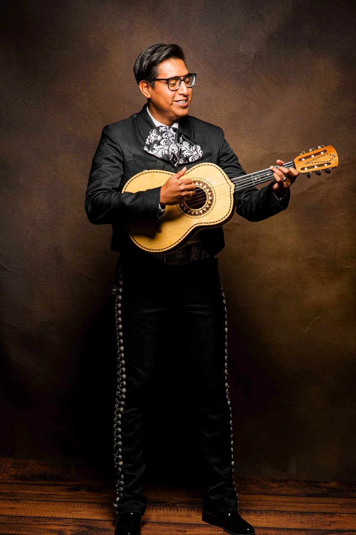 A man dressed in traditional Mexican charro outfit playing a guitar, standing against a dark brown background.