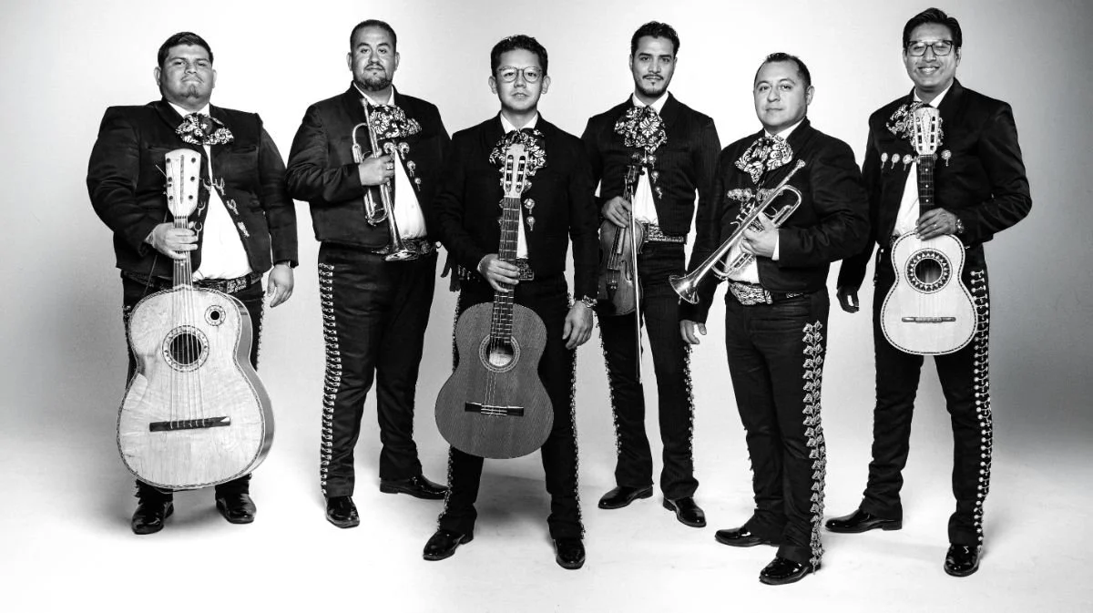 Black and white photo of six musicians dressed in traditional Mexican Mariachi outfits, holding various musical instruments including guitars, a trumpet, and a violin, standing against a plain background.