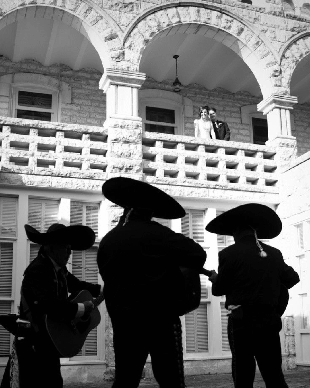 Silhouettes of three mariachi musicians playing guitar and singing, with a couple in wedding attire standing on an upper balcony of a stone building in the background.