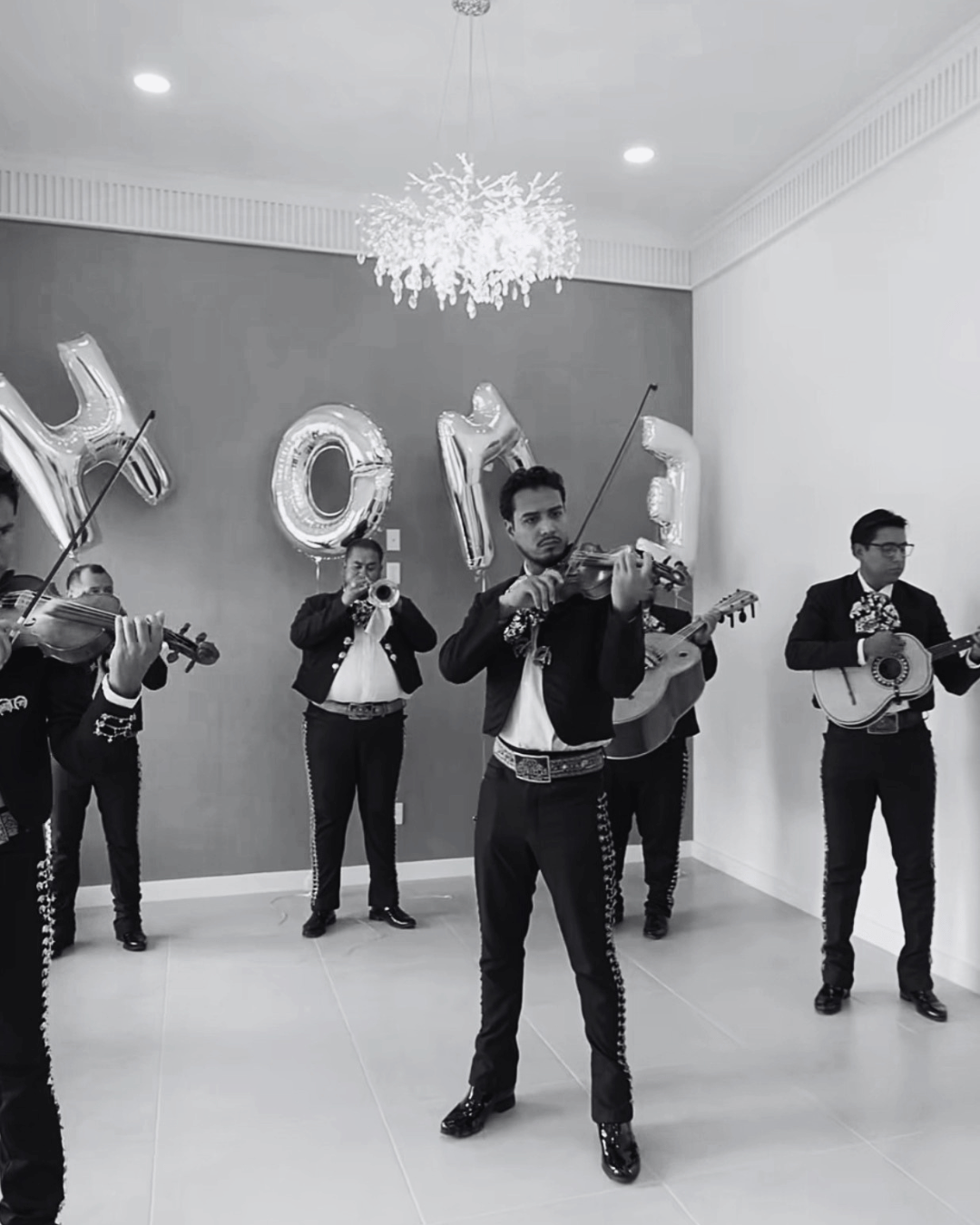A group of musicians playing instruments, including violins, guitar, and trumpet, during a celebration with large balloons spelling 'HOPE' and a chandelier in the background.