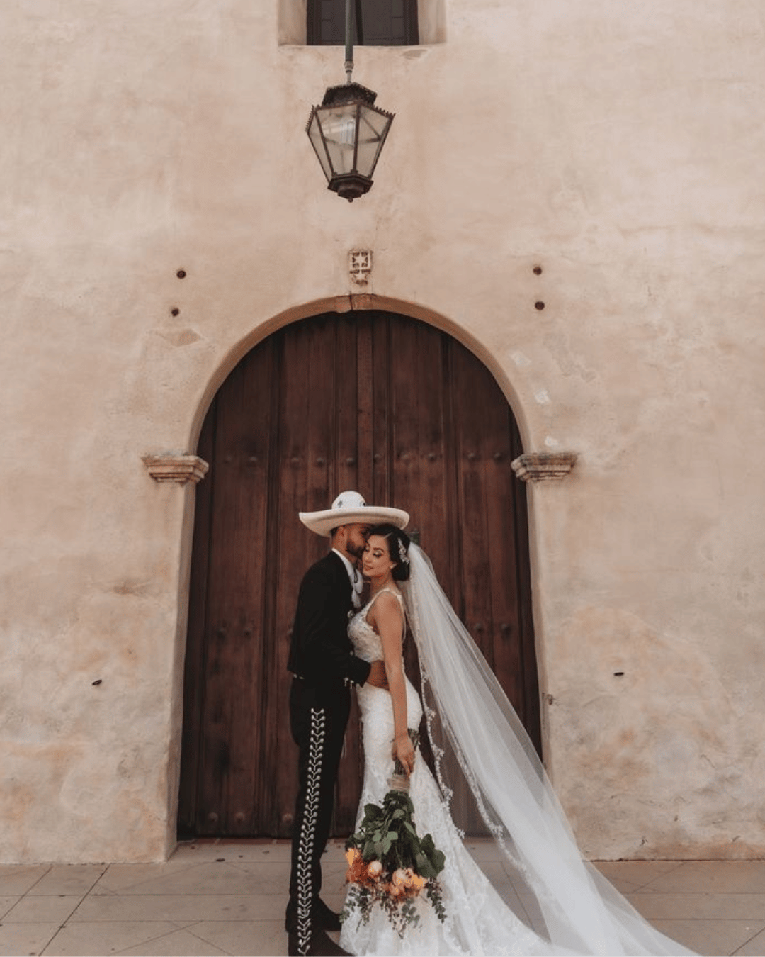 A bride and groom in wedding attire, with the groom wearing a traditional Mexican sombrero, standing close together in front of a large wooden door on a textured beige wall, behind them a vintage lantern hangs from the wall.