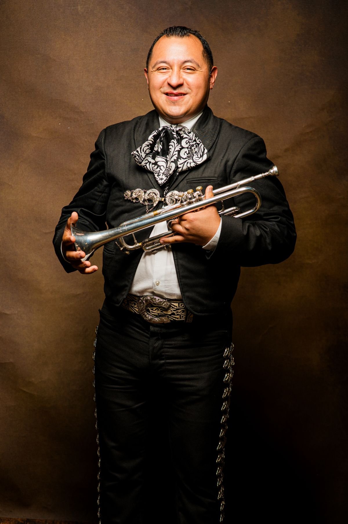 Man dressed in traditional Mexican mariachi attire holding a trumpet, smiling, against a brown background.