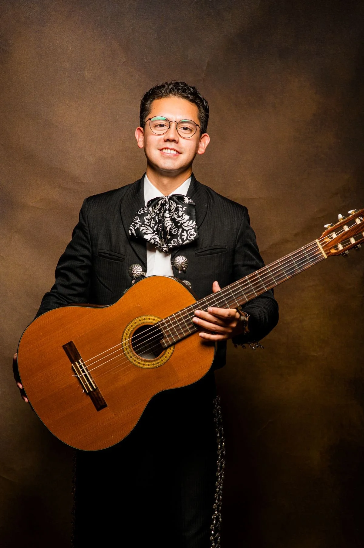 A man dressed in traditional Mexican charro attire, holding a guitar, standing in front of a brown background.