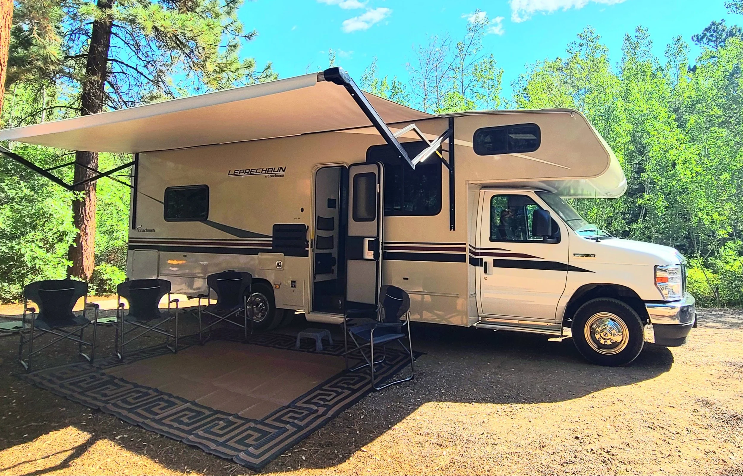 A white camper van with a retractable awning set up underneath tall green trees on a bright sunny day. There are black chairs arranged around a small table outside the camper. The ground is dirt and a patterned rug is laid out beneath the chairs.