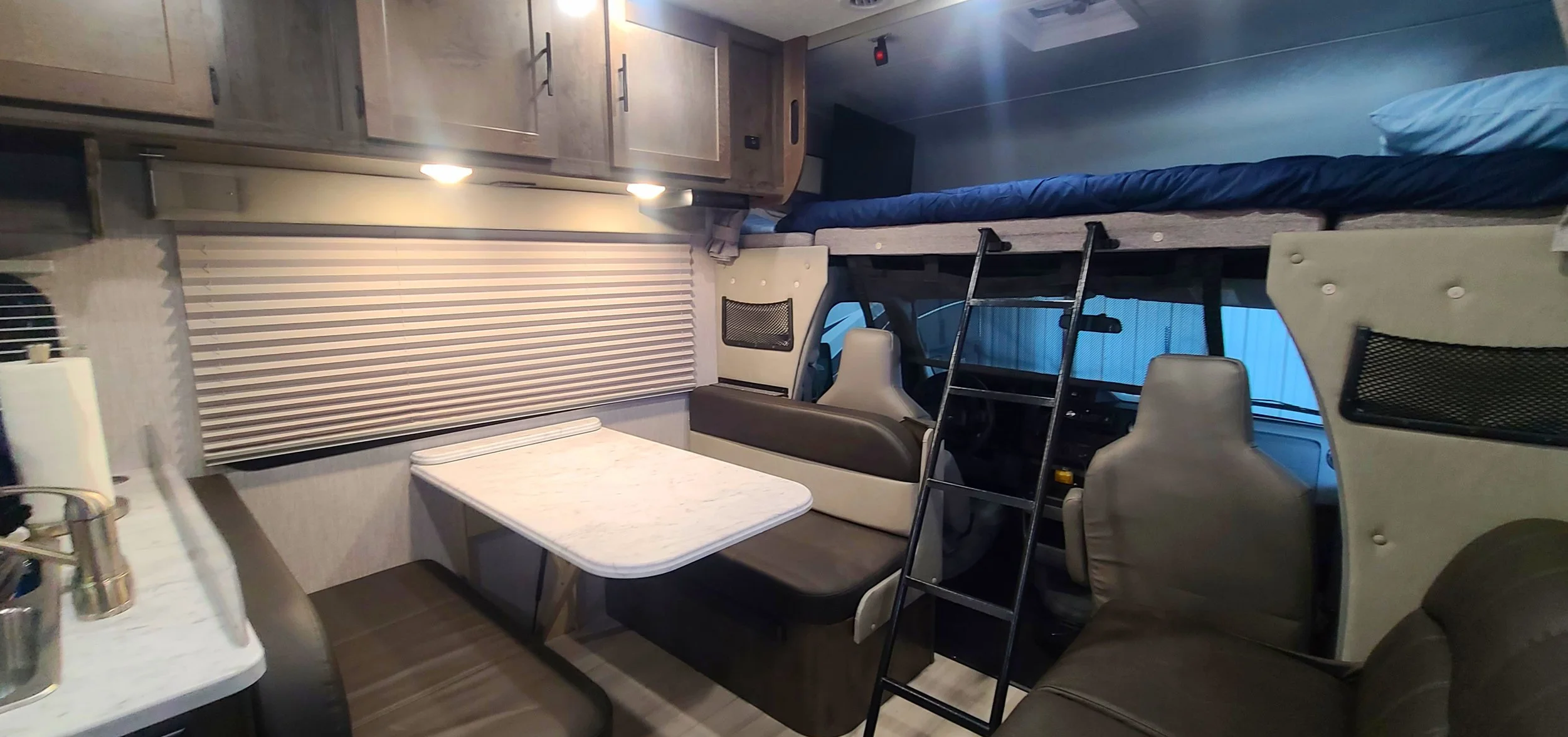 Interior of a recreational vehicle with a small dining area, a bed above the driver's cabin, and various furniture, including chairs and a table.