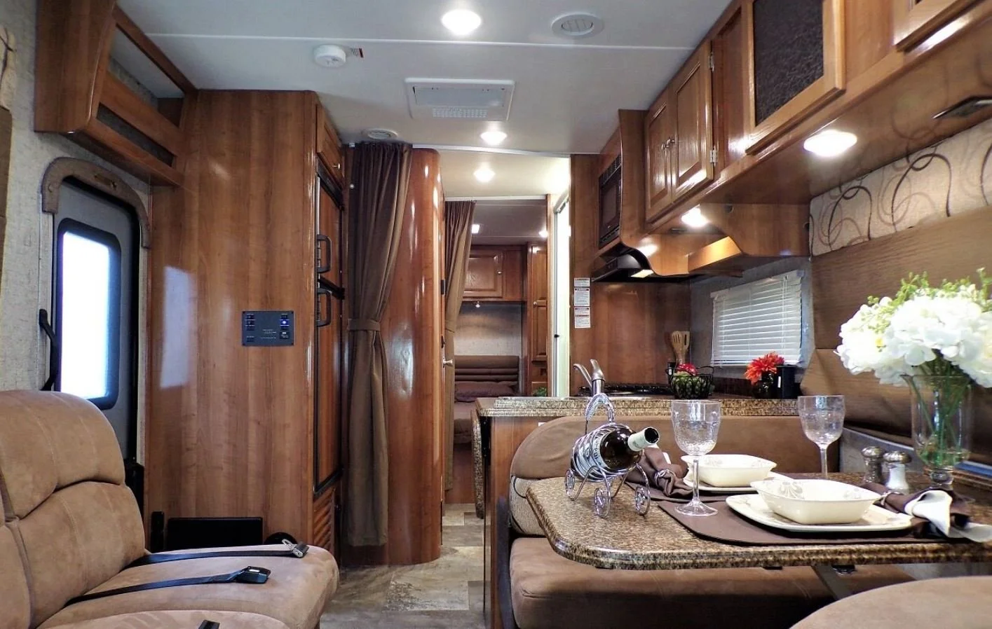 Interior of an RV with a dining table set with wine glasses, bowls, and a flower arrangement, and a kitchen area with wooden cabinets and a window with blinds.