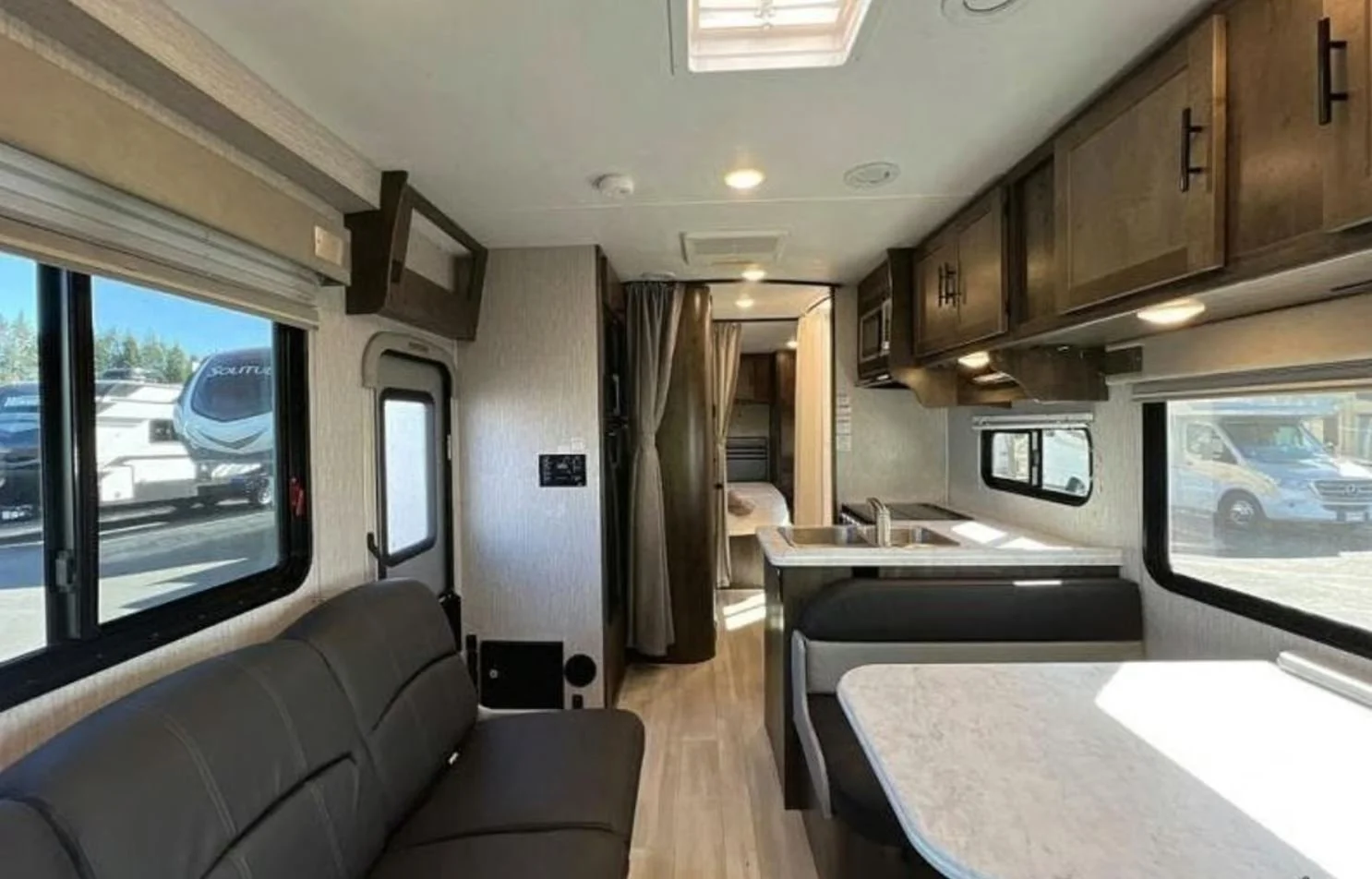 Interior of an RV with black sofa, dining table, kitchen area with sink and cabinets, and a partition with curtains leading to a sleeping area with bed.