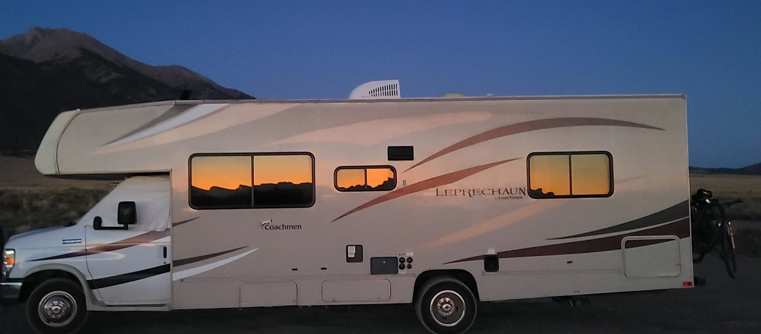 A beige and brown Coachmen Leprechaun campervan parked outdoors at dusk, with a mountain in the background and sunset reflections in the windows.