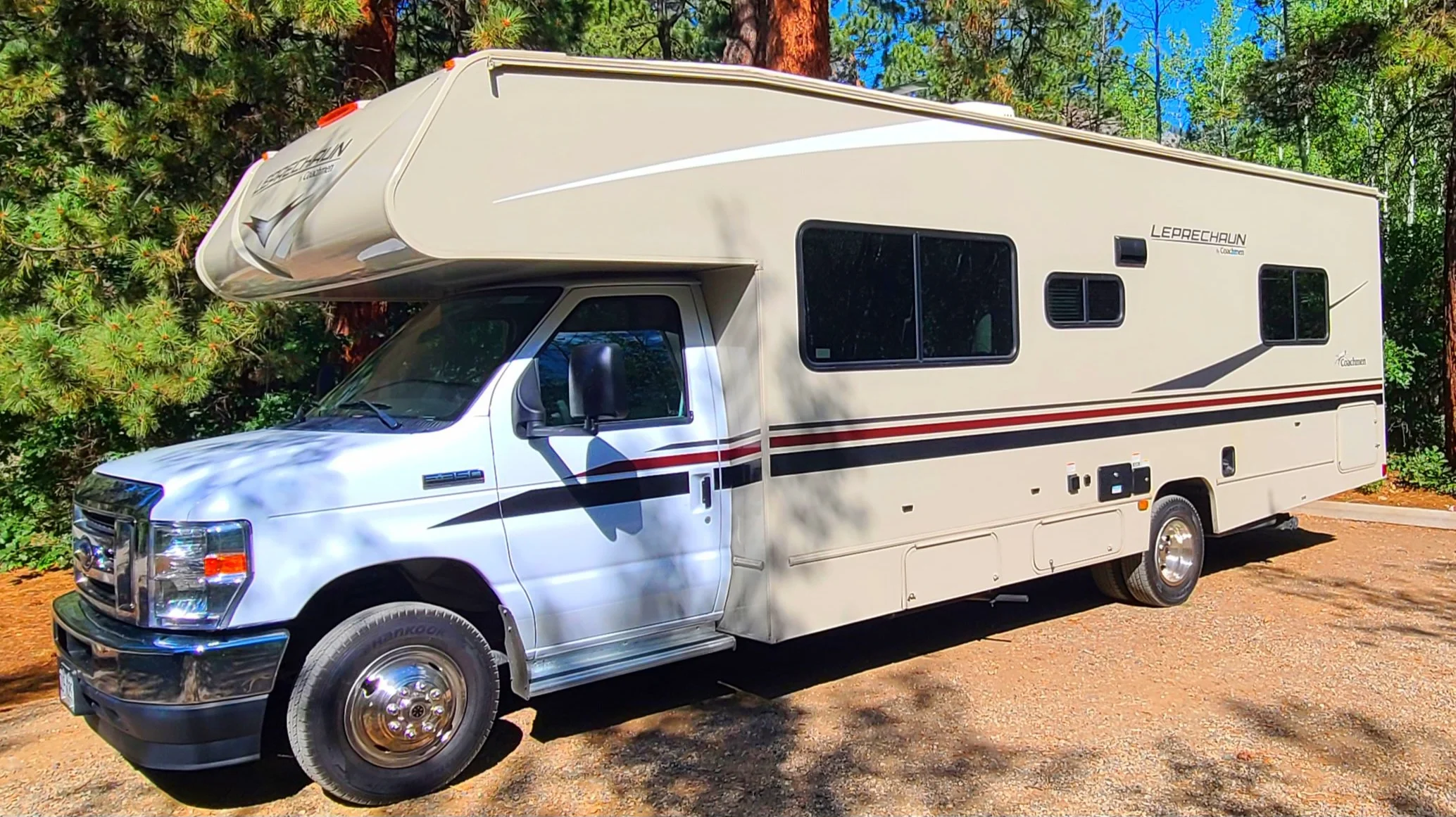 A white camper van with black and red stripes parked on dirt ground amidst green pine trees and tall mountains in the background.