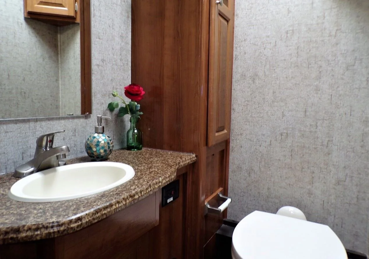 Bathroom with granite countertop, white sink, wooden cabinets, mirror, soap dispenser, a vase with a red rose, and a toilet.