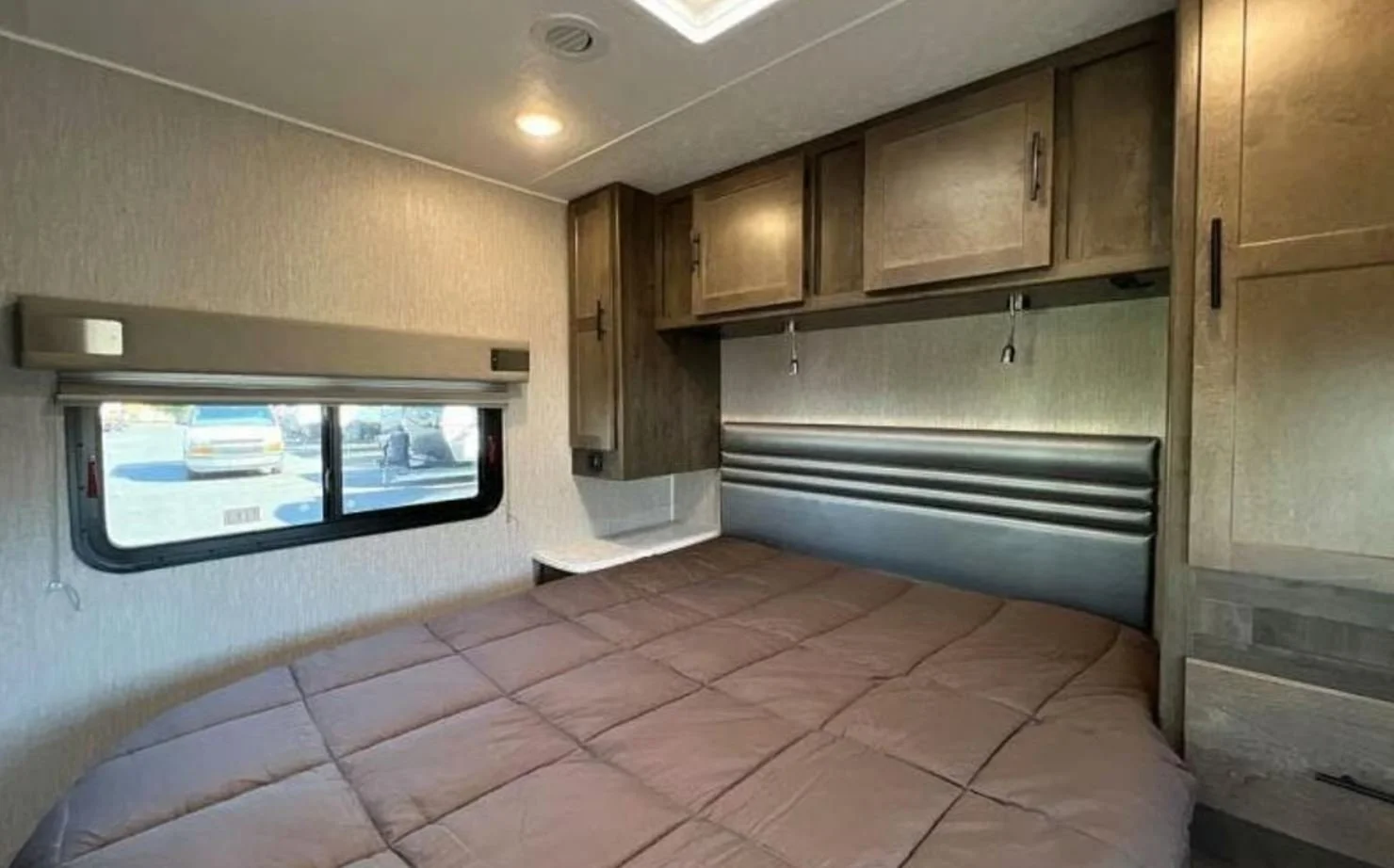 Interior of a bedroom in an RV or camper with a bed, overhead cabinets, a window to the outside, and a small nightstand.