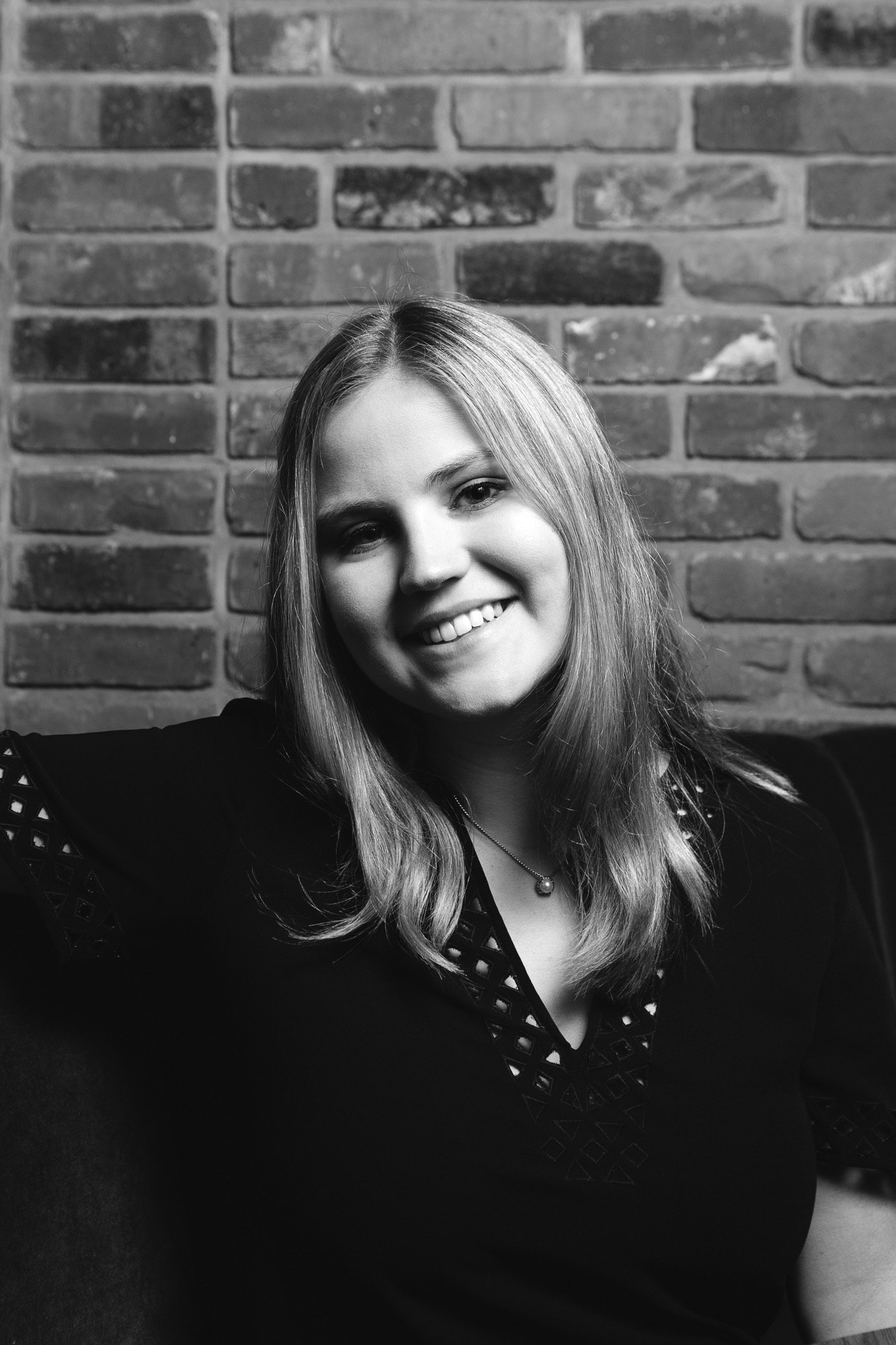 Close-up of a smiling woman with long hair, wearing a black top with cut-out designs, against a brick wall background.