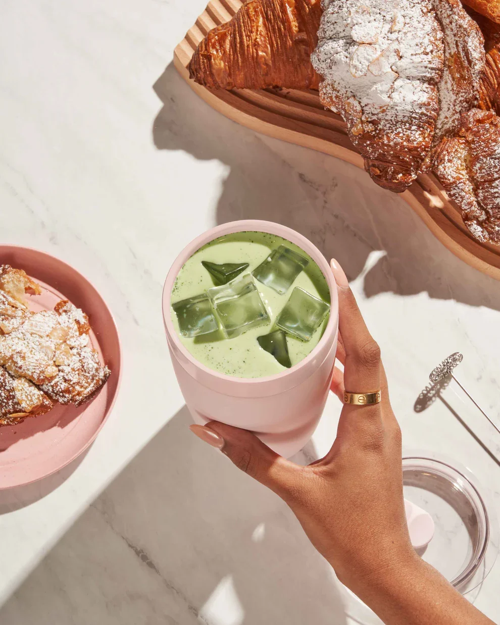 A hand holding a pink cup of iced matcha latte with ice cubes and mint leaves, placed on a white marble surface. In the background, there are two plates with croissants dusted with powdered sugar, one on a pink plate and the other on a circular wooden tray.