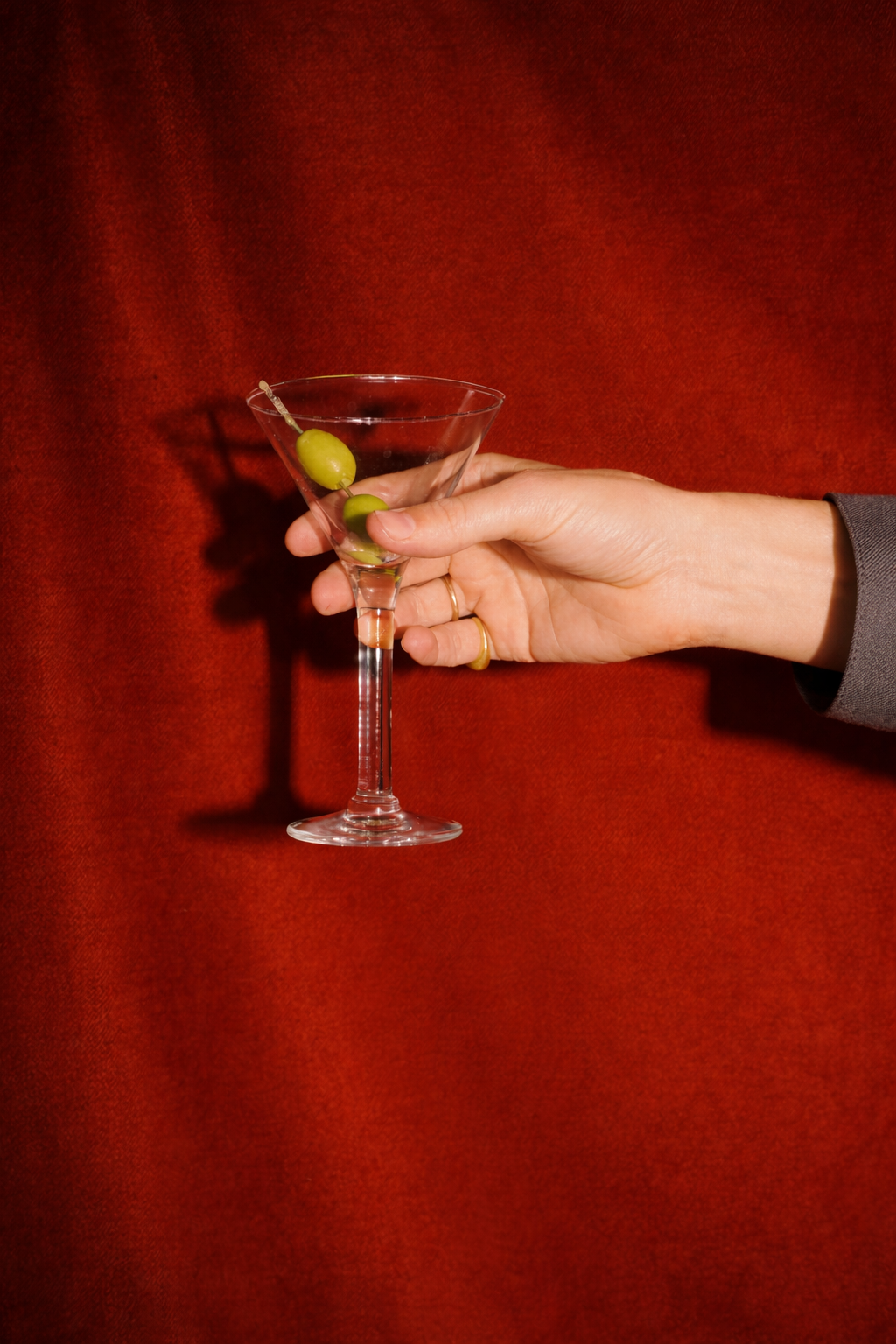 A hand holding a martini glass with green olives on a skewer against a red background.