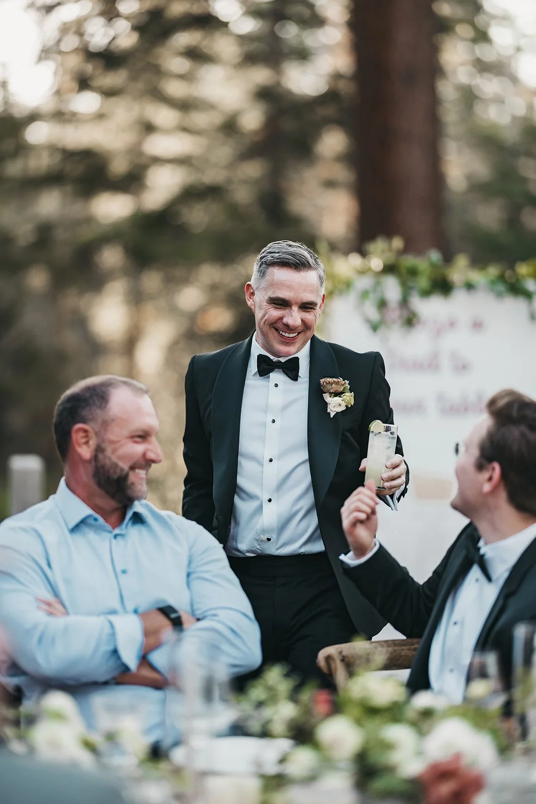 A man in a tuxedo is smiling and holding a drink, standing outdoors at a wedding reception with two seated men, one in a light blue shirt and one in a tuxedo, all enjoying a conversation.