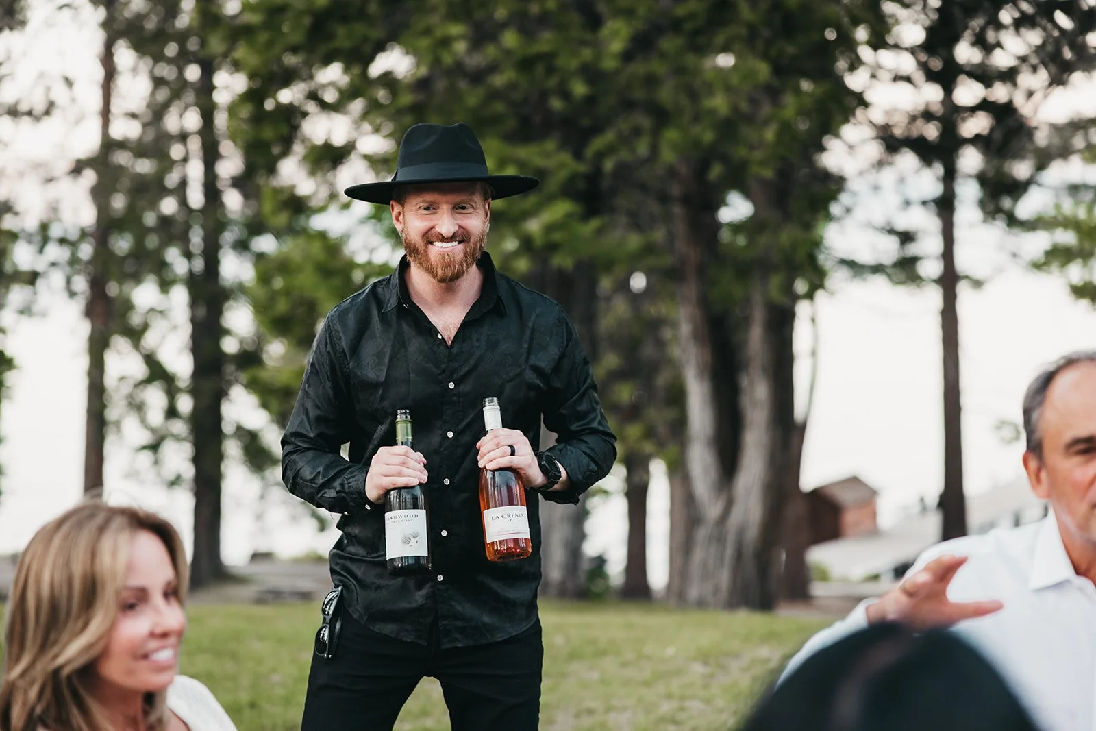 A man with a beard wearing a black hat and black shirt stands outdoors holding a bottle of red wine and a bottle of white wine. He smiles as he looks at someone. There are people sitting nearby, and trees are in the background.