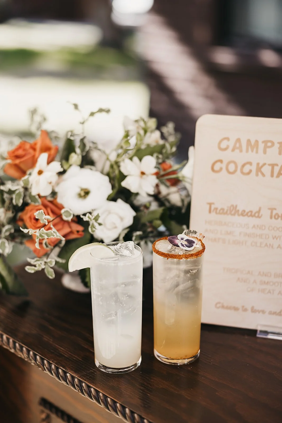 Two cocktails, one clear with ice and a lemon slice, the other yellow with a floral garnish, placed on a wooden surface with a floral arrangement and a sign in the background.