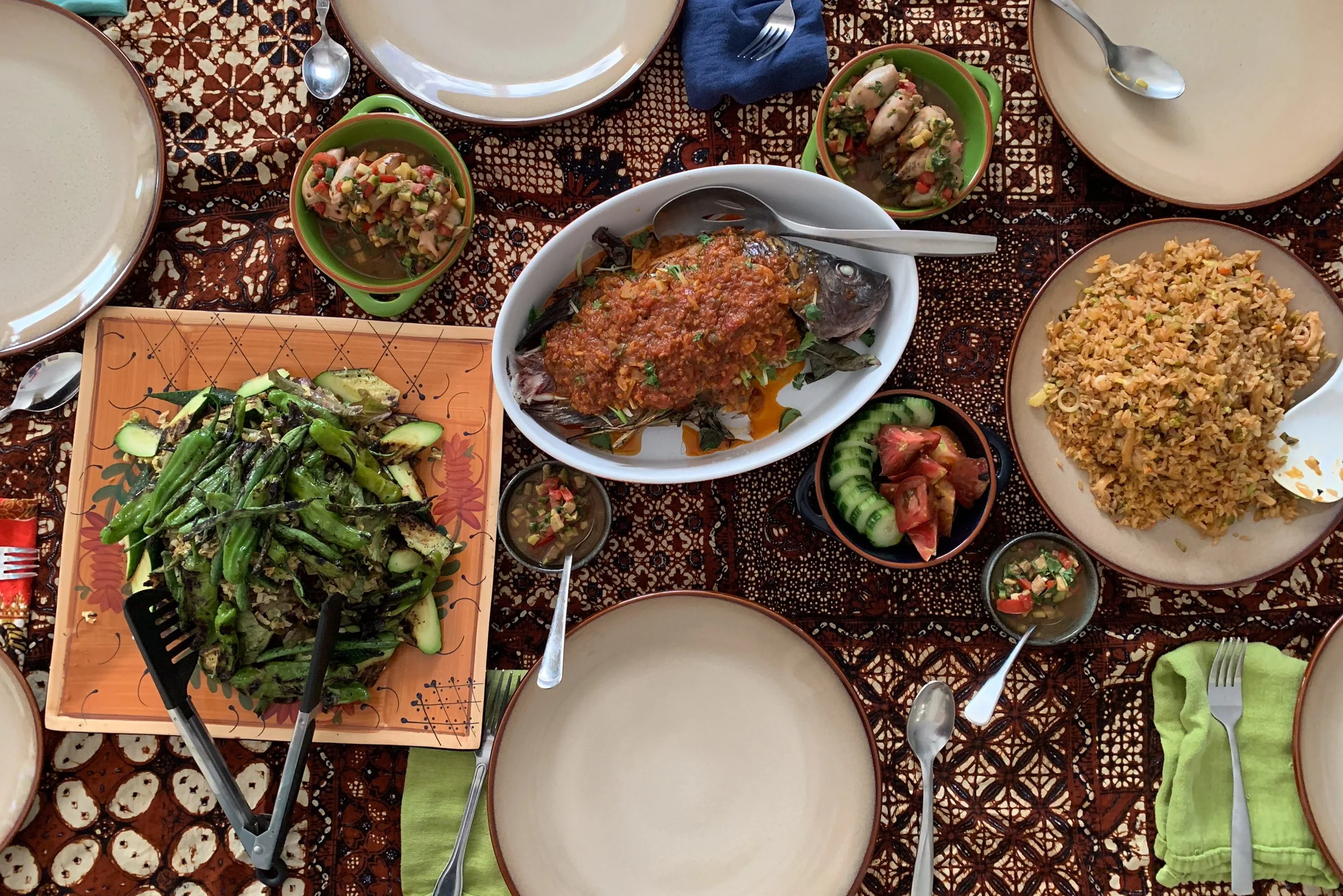 A table set for a meal with empty plates, various bowls of food including a whole fish topped with sauce, rice, salad, and small cups of sauce. There are utensils, napkins, and a patterned tablecloth.