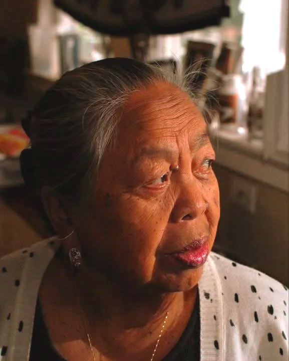 Close-up of an elderly Asian woman with gray hair tied back, looking out a window with a contemplative expression.