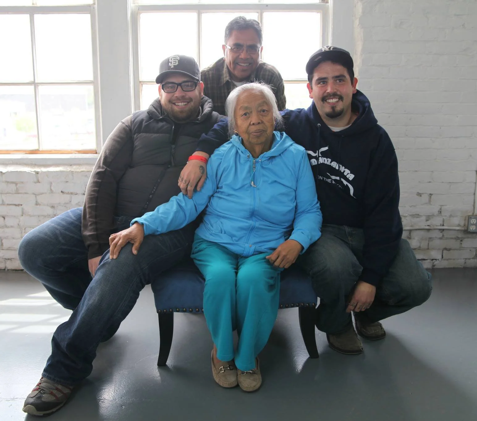 A group of five people, four men and one elderly woman, sitting and standing in front of a large window with white brick walls behind them. They are smiling and appear to be posing for a photo.