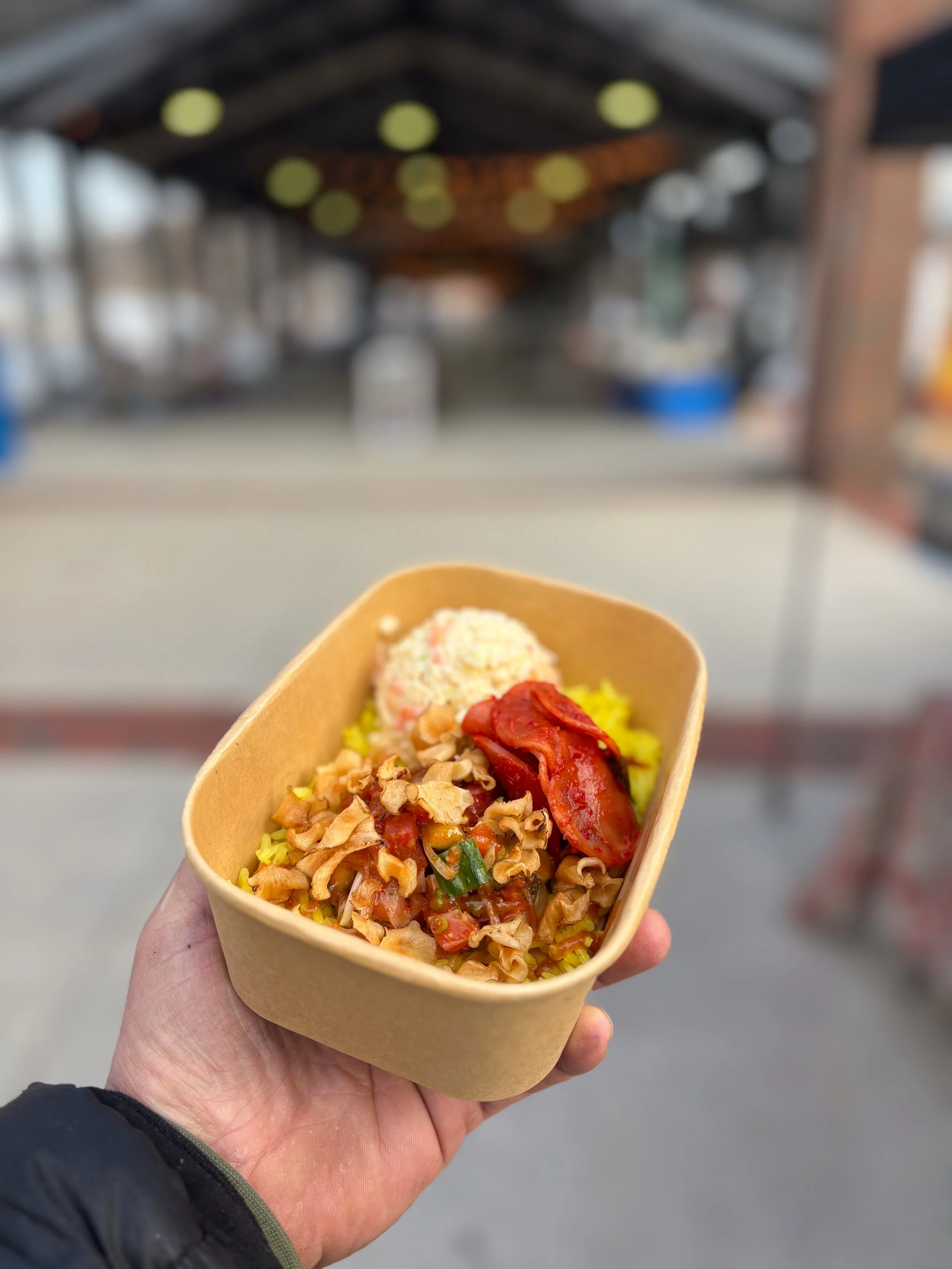 A person holding a brown paper food container filled with fried rice, topped with sliced red meats, vegetables, and possibly some grilled meat, with a blurred outdoor market or street scene in the background.