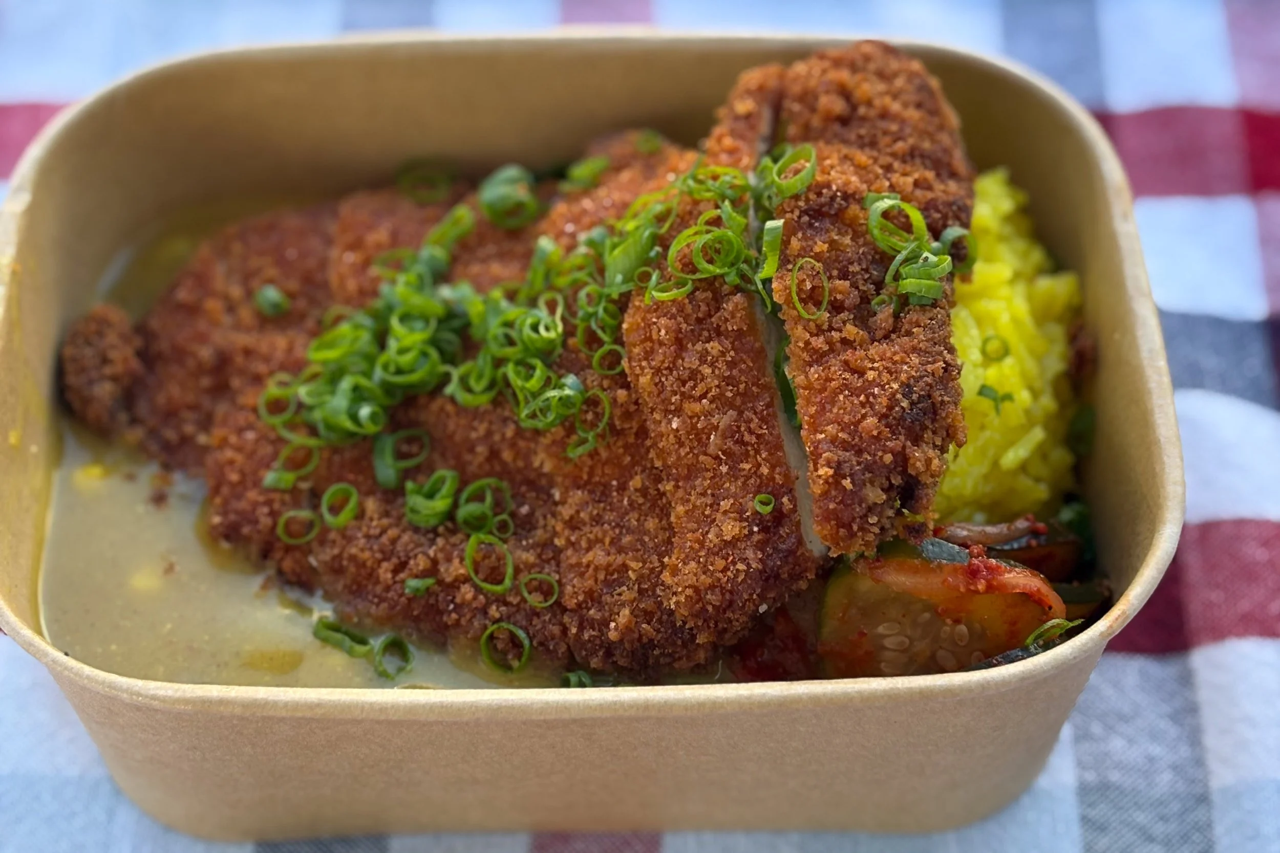 Fried chicken katsu topped with green onions served over rice and curry in a takeout container.