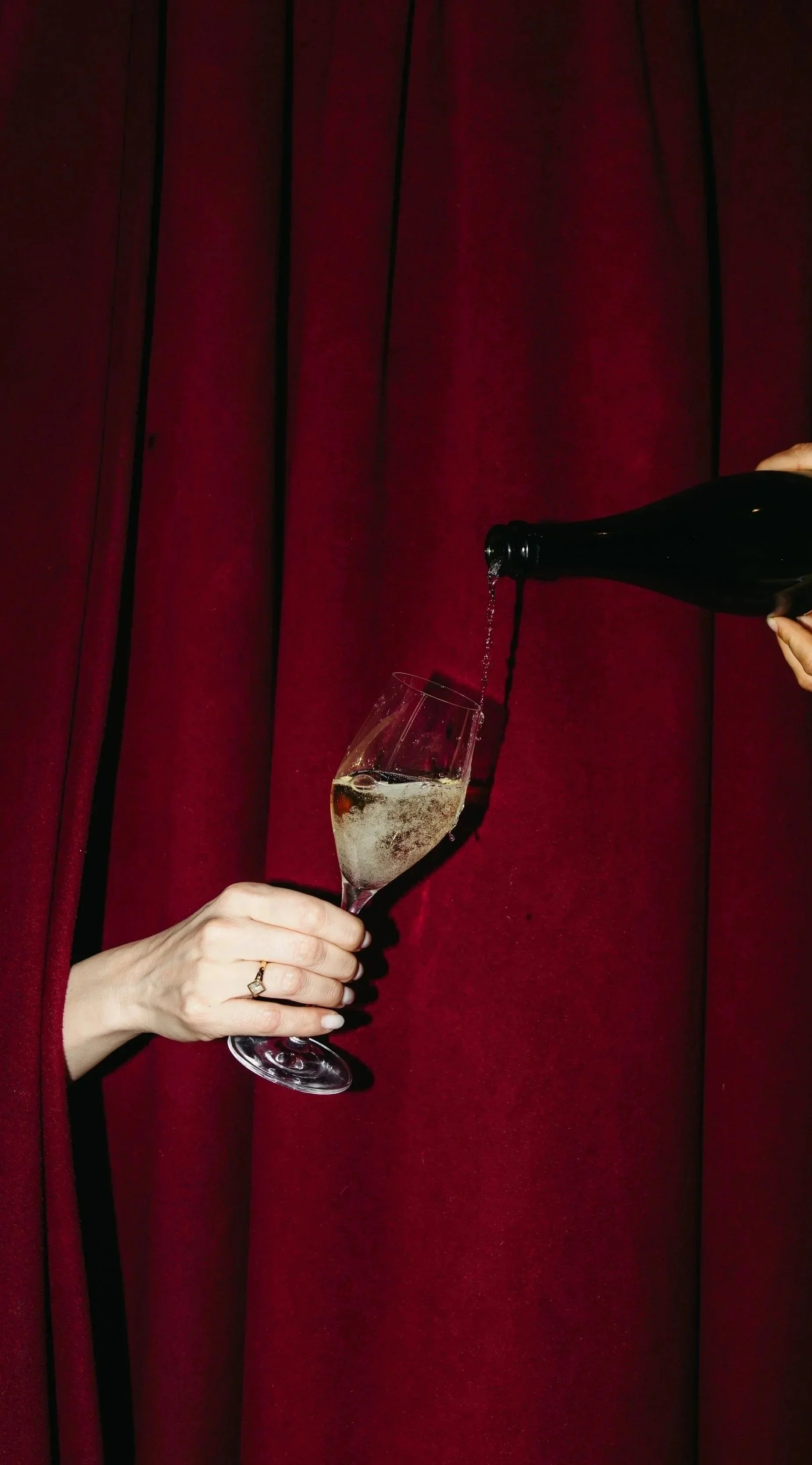 Two hands — one holding a glass, one pouring champagne — framed by a red curtain