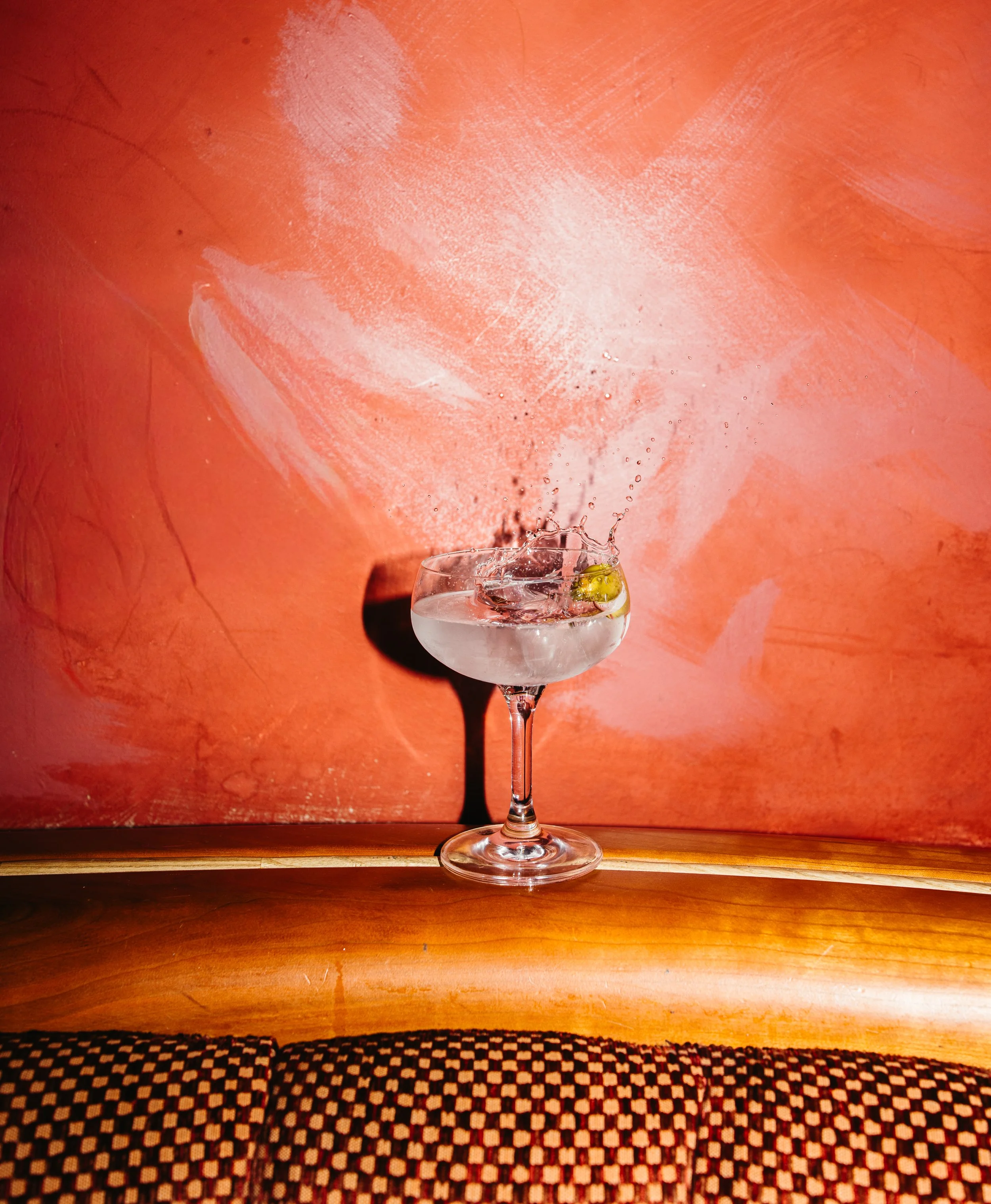 A wine glass filled with ice and a lime wedge on a wooden surface, with a bright orange painted wall in the background.