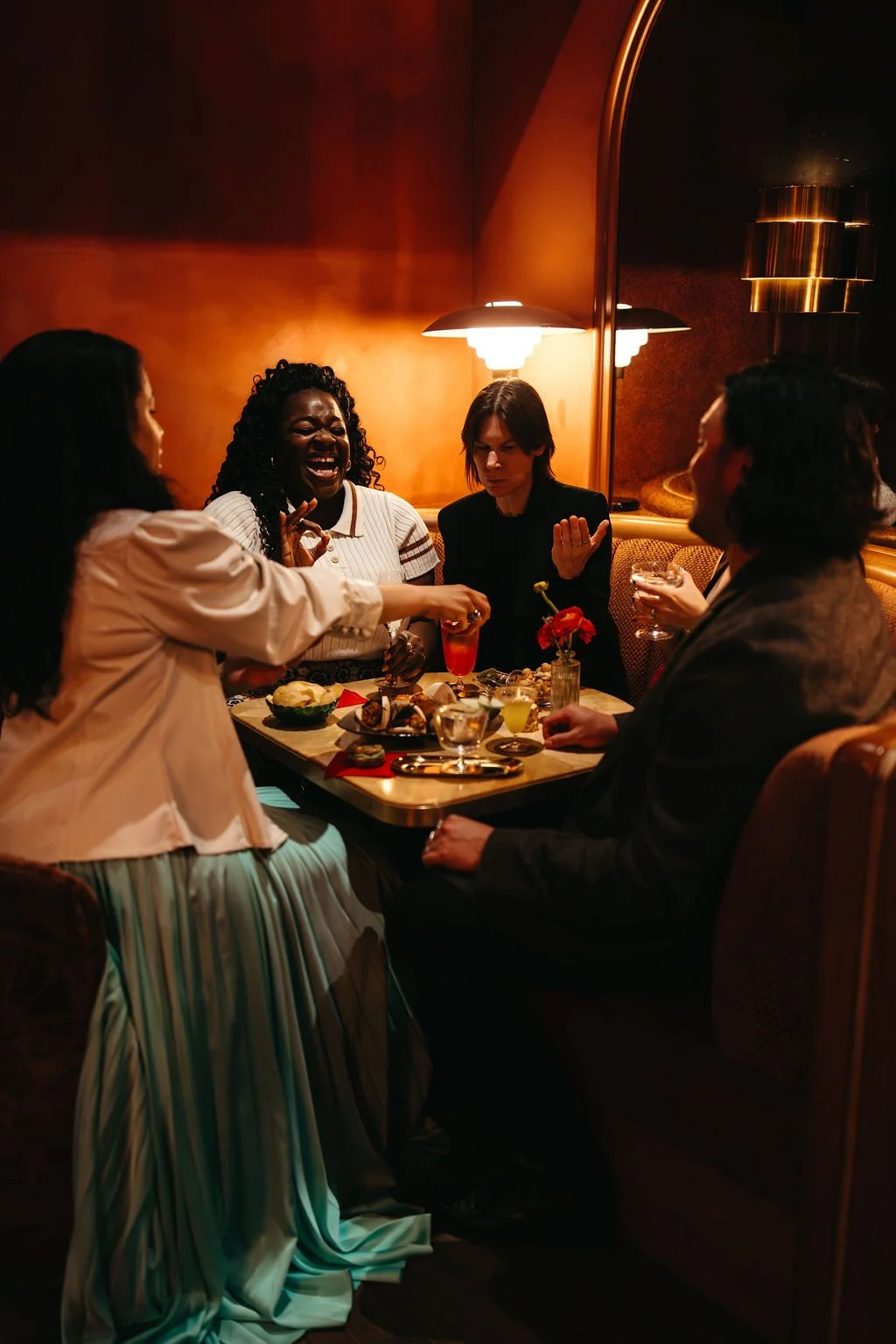 Group of five people laughing and talking at a restaurant table with food and drinks, warmly lit interior.