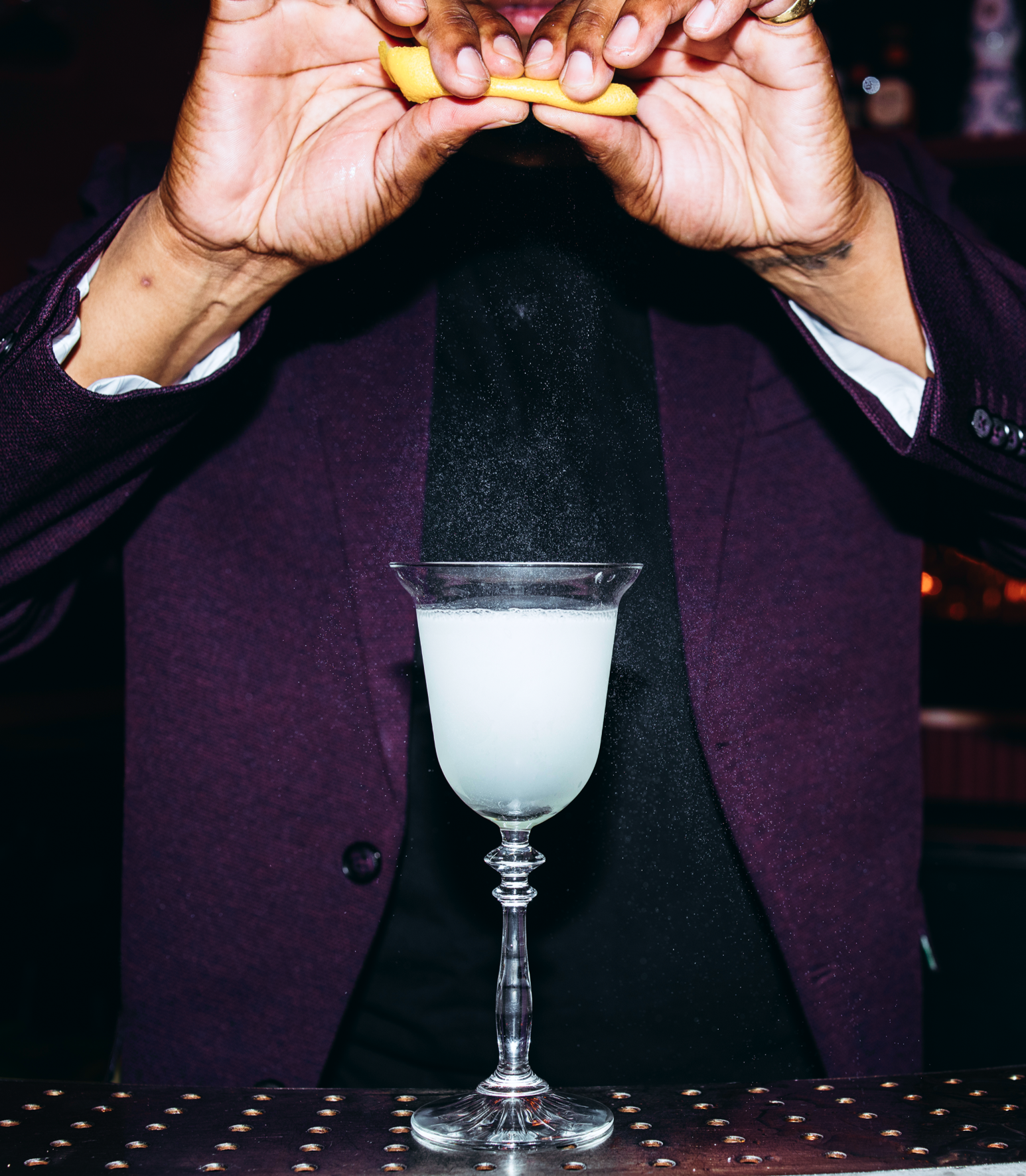 A bartender squeezing a lemon twist over a pouring cocktail, citrus misting in the air