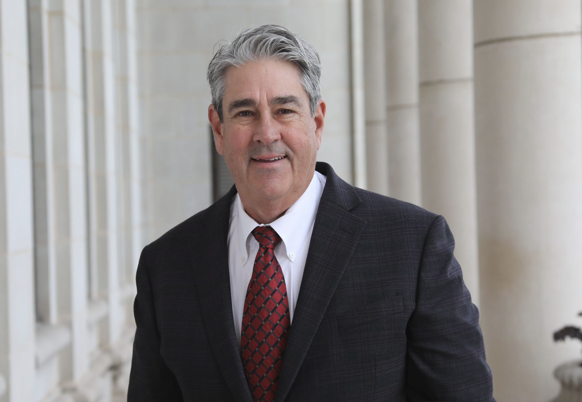 A middle-aged man with gray hair wearing a dark suit, white shirt, and red patterned tie, smiling while walking outdoors in front of a building with large columns.