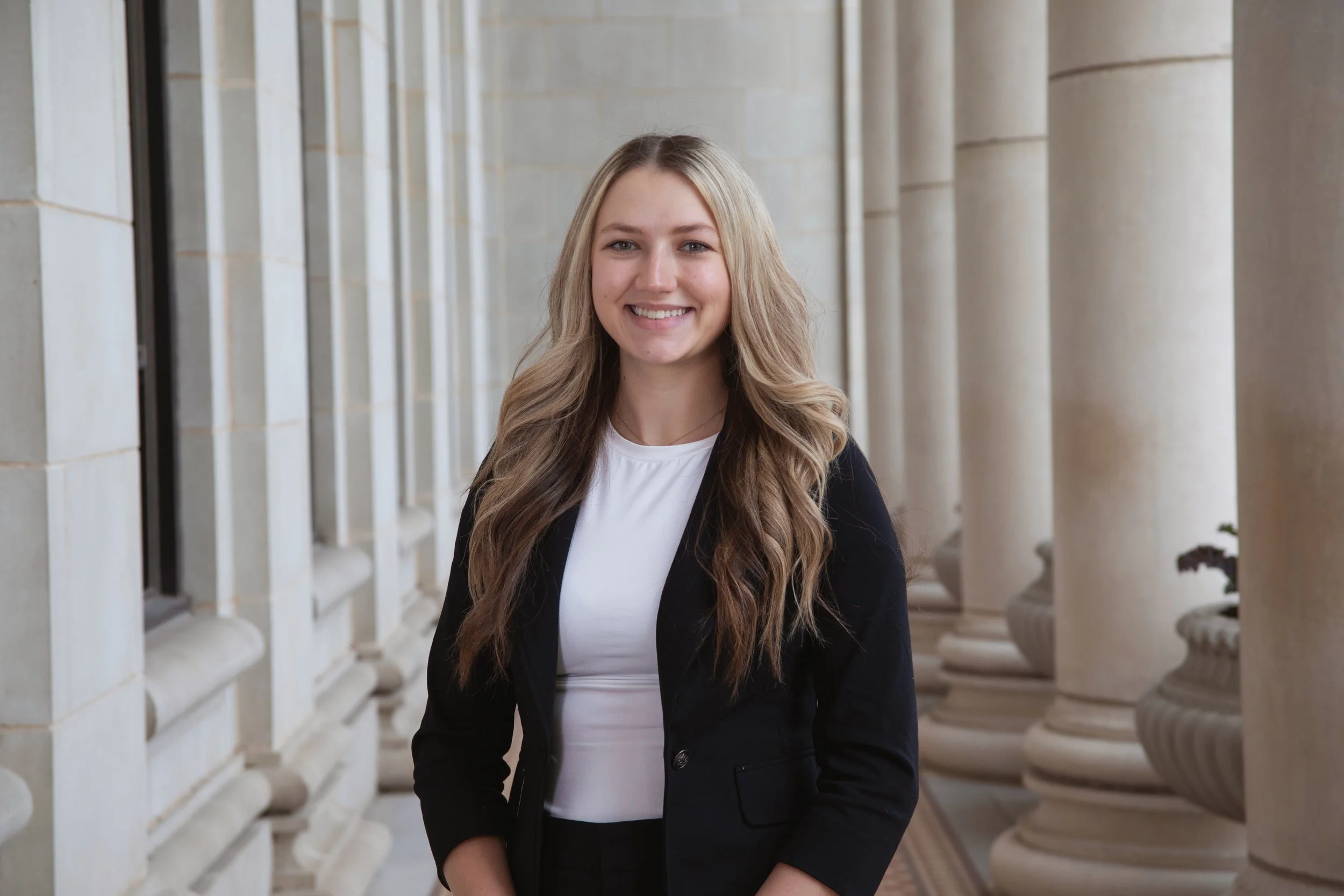 A young woman with long, wavy blonde hair and a bright smile, standing in front of a row of white classical columns.