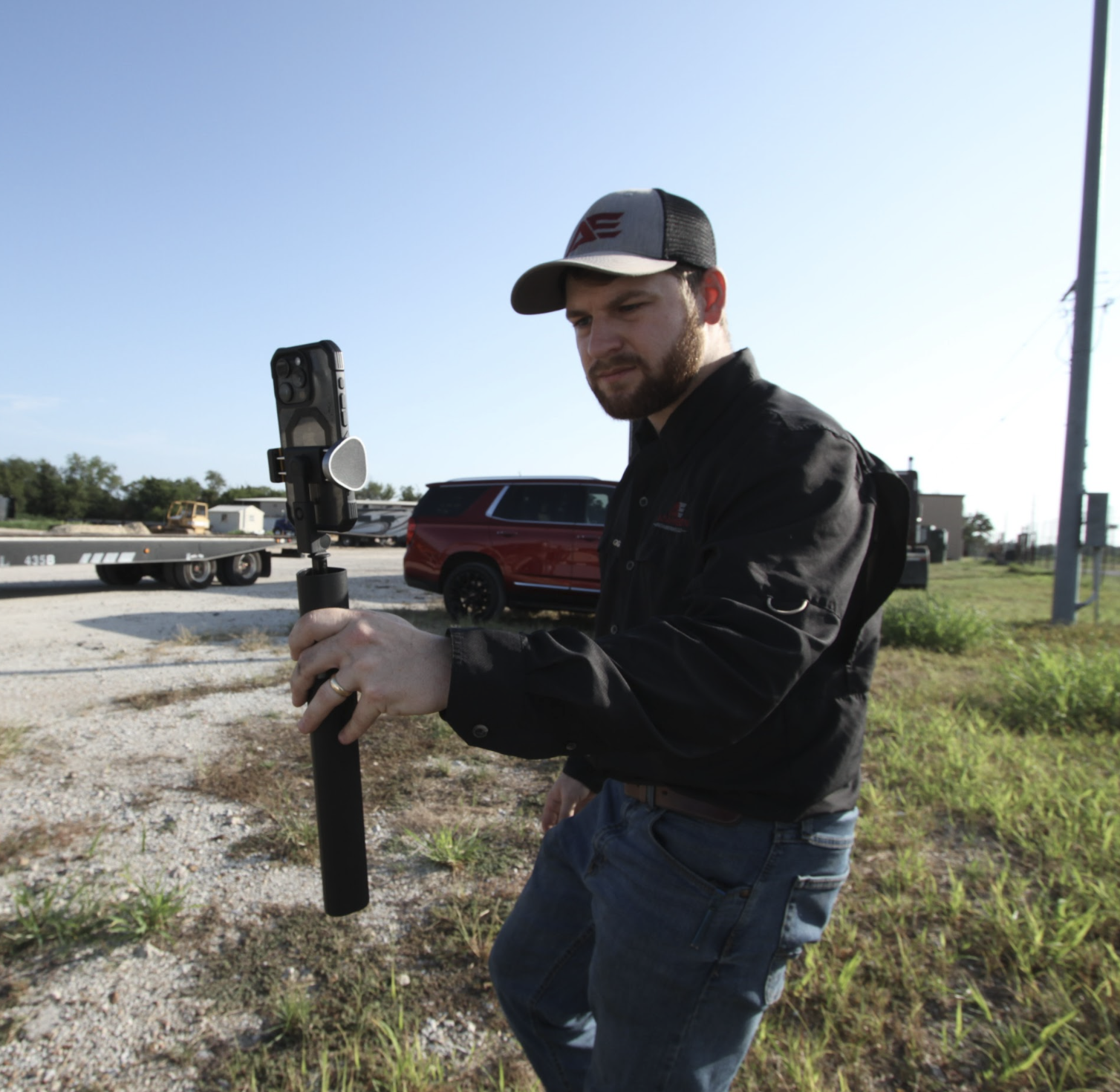 A man wearing a black shirt and a gray and black cap is standing outdoors in a grassy area. He is holding a camera stabilizer mounted on a pole and appears to be looking at the camera screen. In the background, there are vehicles, including a red SUV, a trailer, and a few buildings under a clear blue sky.