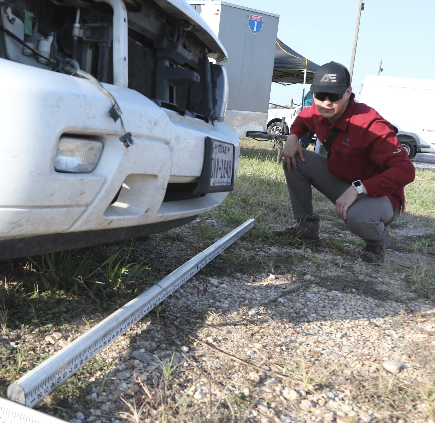 A man dressed in a red jacket, gray pants, black hat, and sunglasses, crouching next to the front of a white vehicle with its hood open, on a gravel area with a measuring scale on the ground.