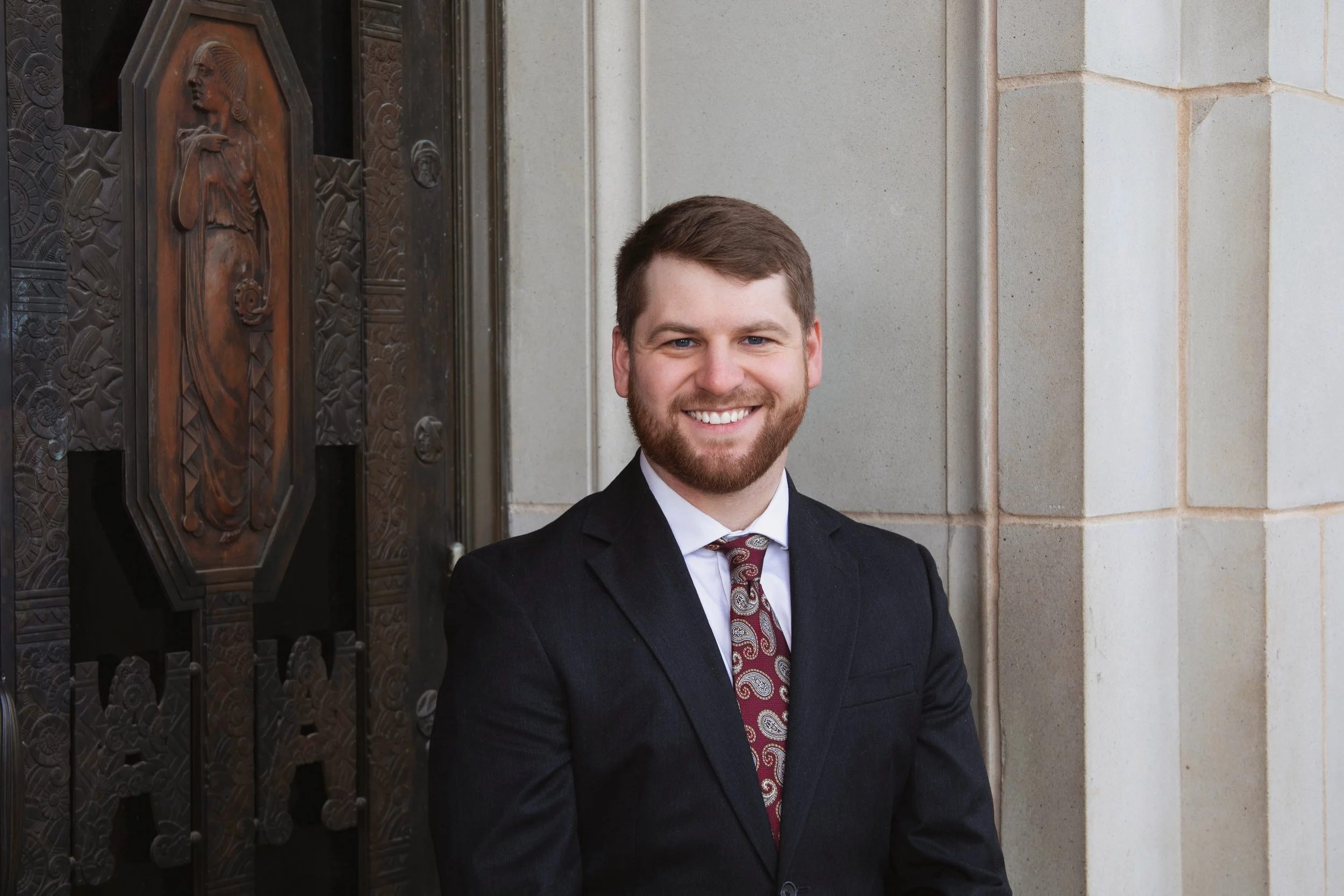 A smiling man with a beard and short brown hair, wearing a dark suit, white shirt, and patterned tie, standing outdoors in front of a decorative metal door and stone wall.