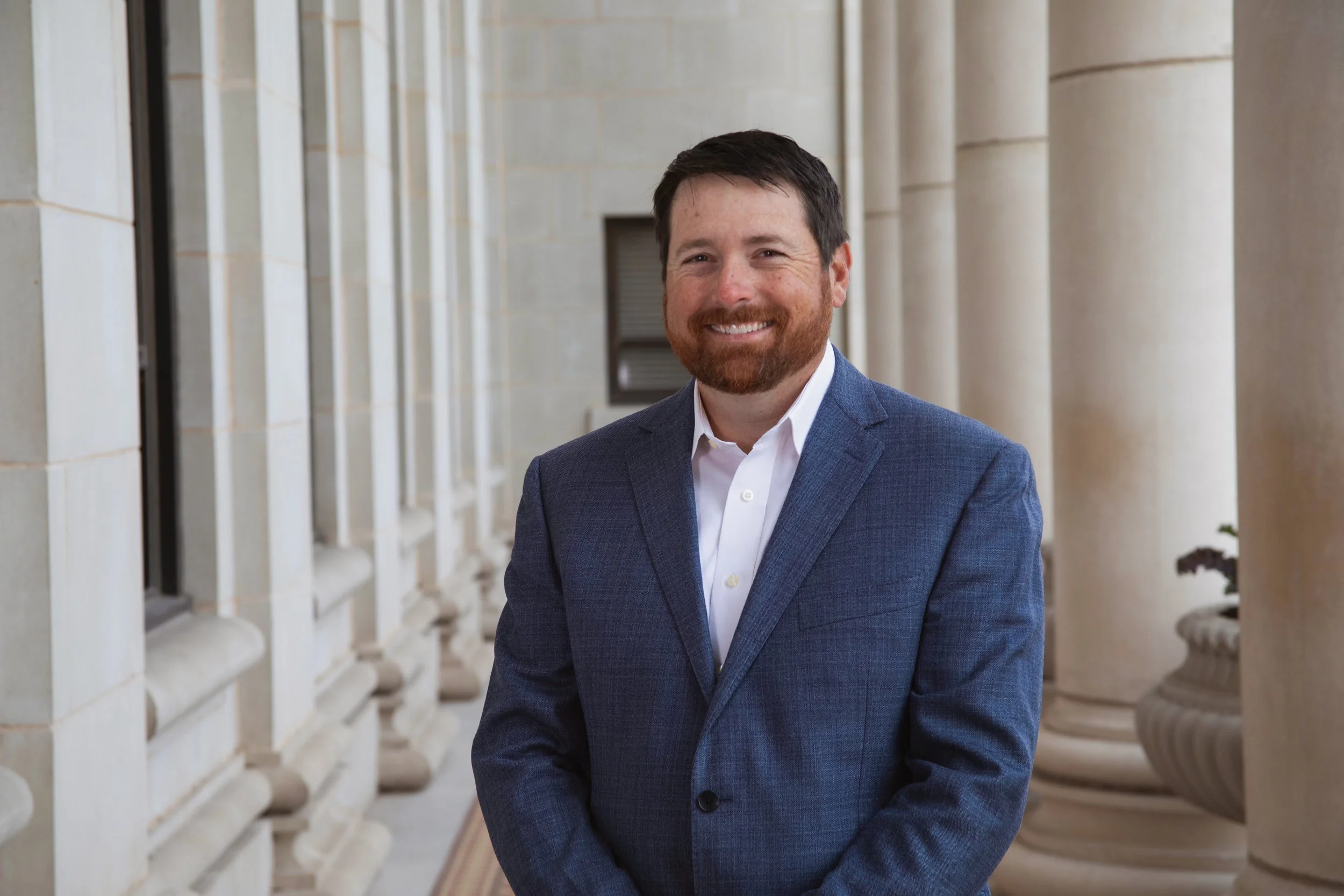 A man with dark hair and a beard smiling in front of stone columns and architectural details in a classical building.