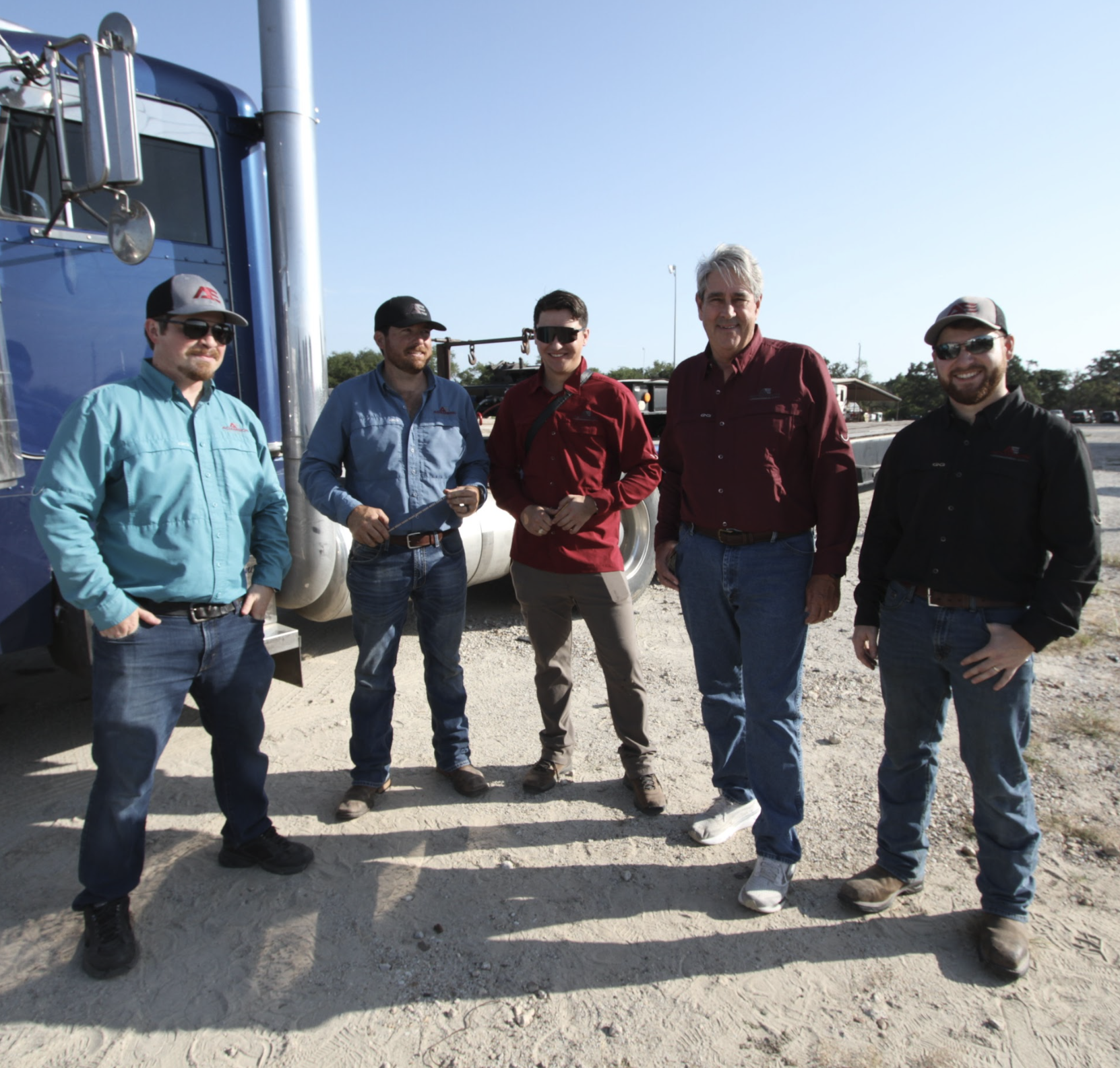 Five men standing outdoors on a gravel surface near a large blue truck with a clear blue sky in the background.