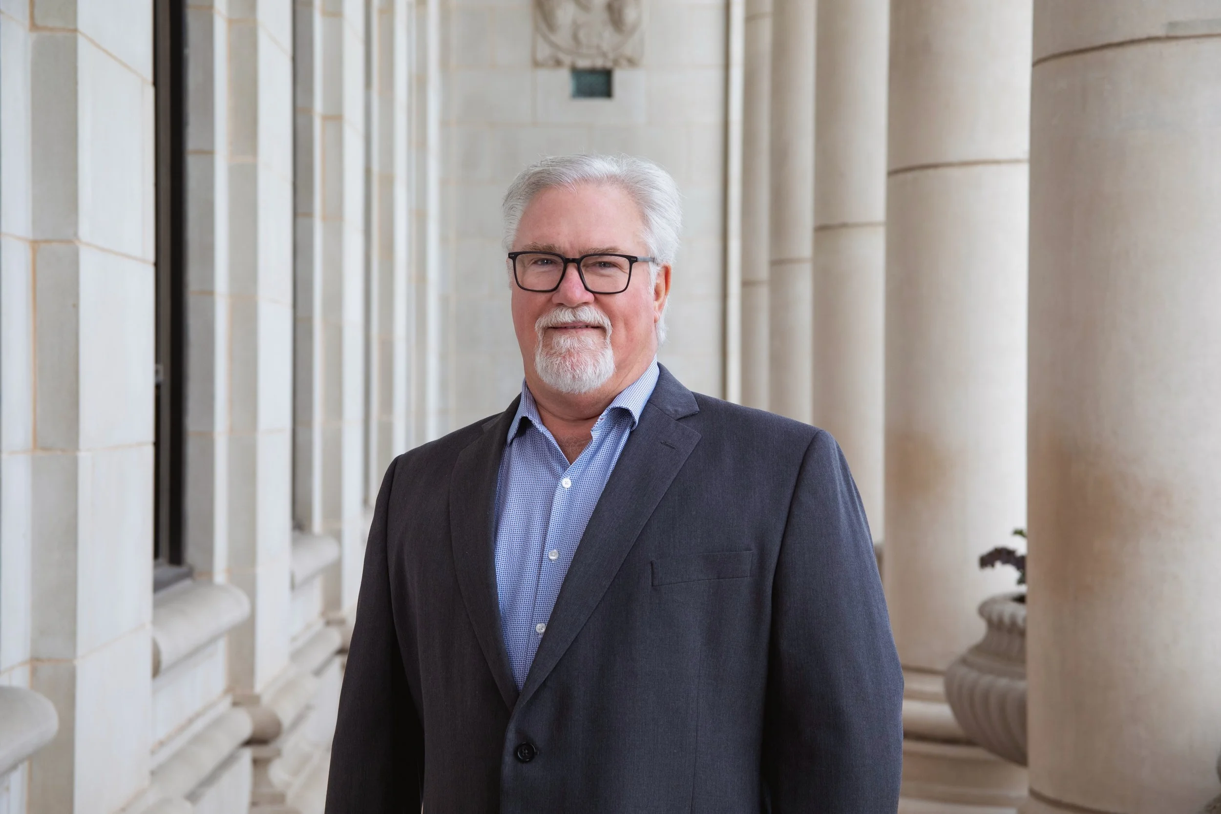 A middle-aged man with gray hair, glasses, and a white beard, wearing a dark suit jacket and a blue collared shirt, standing in front of a beige stone building with columns.
