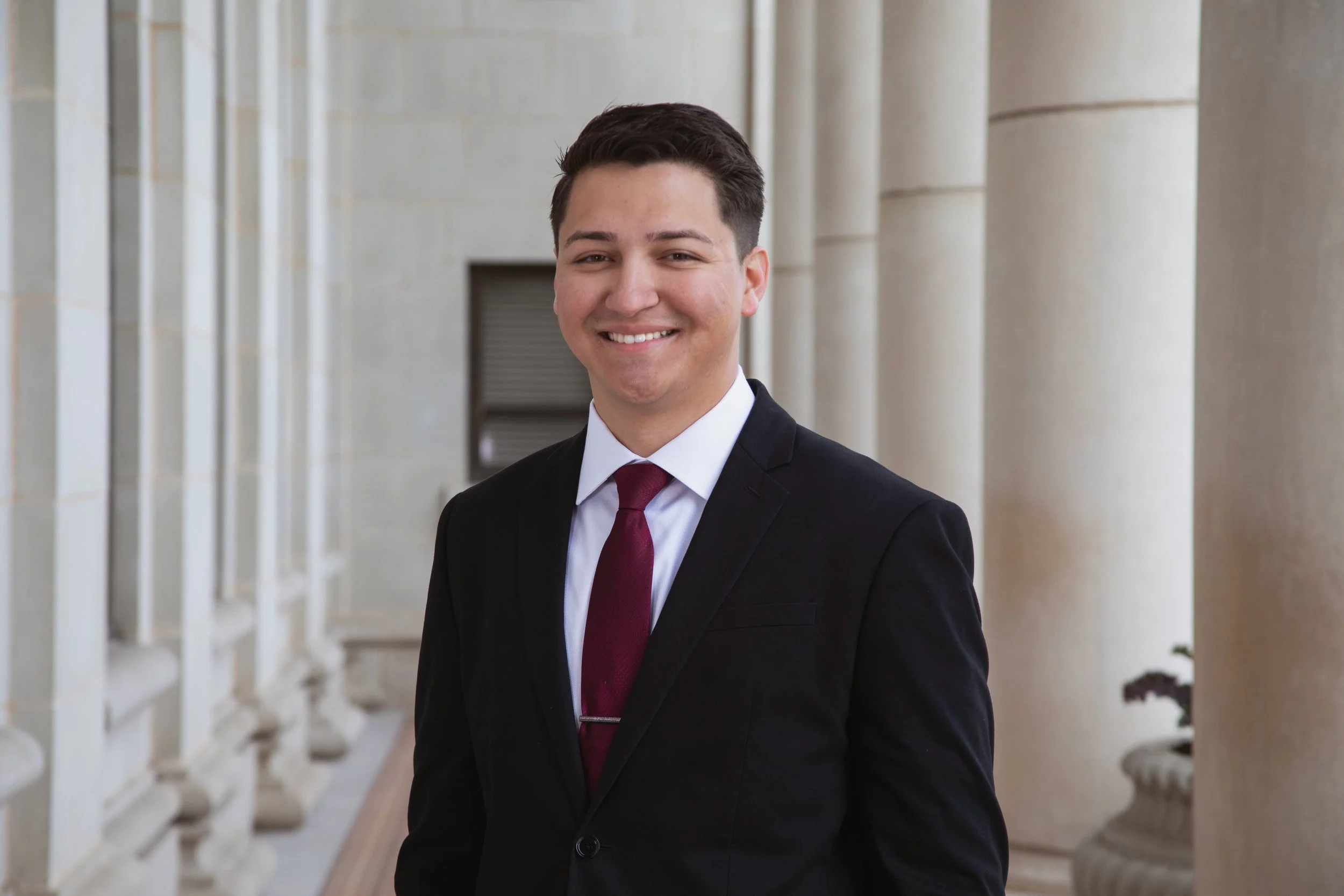 Young man in a suit smiling outdoors near architectural columns.