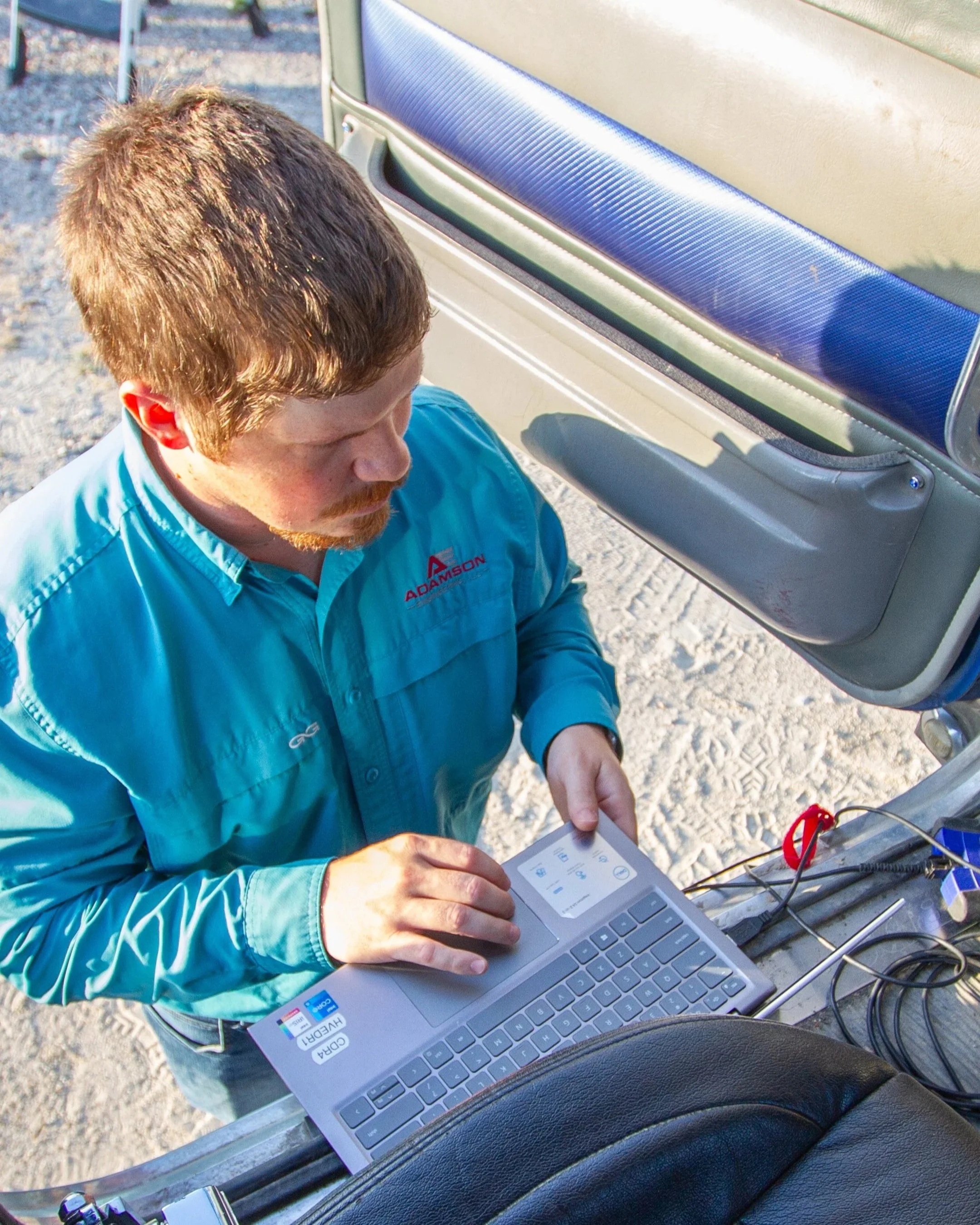 Man with reddish hair and beard using a laptop with various equipment, sitting outdoors near a vehicle with an open door.