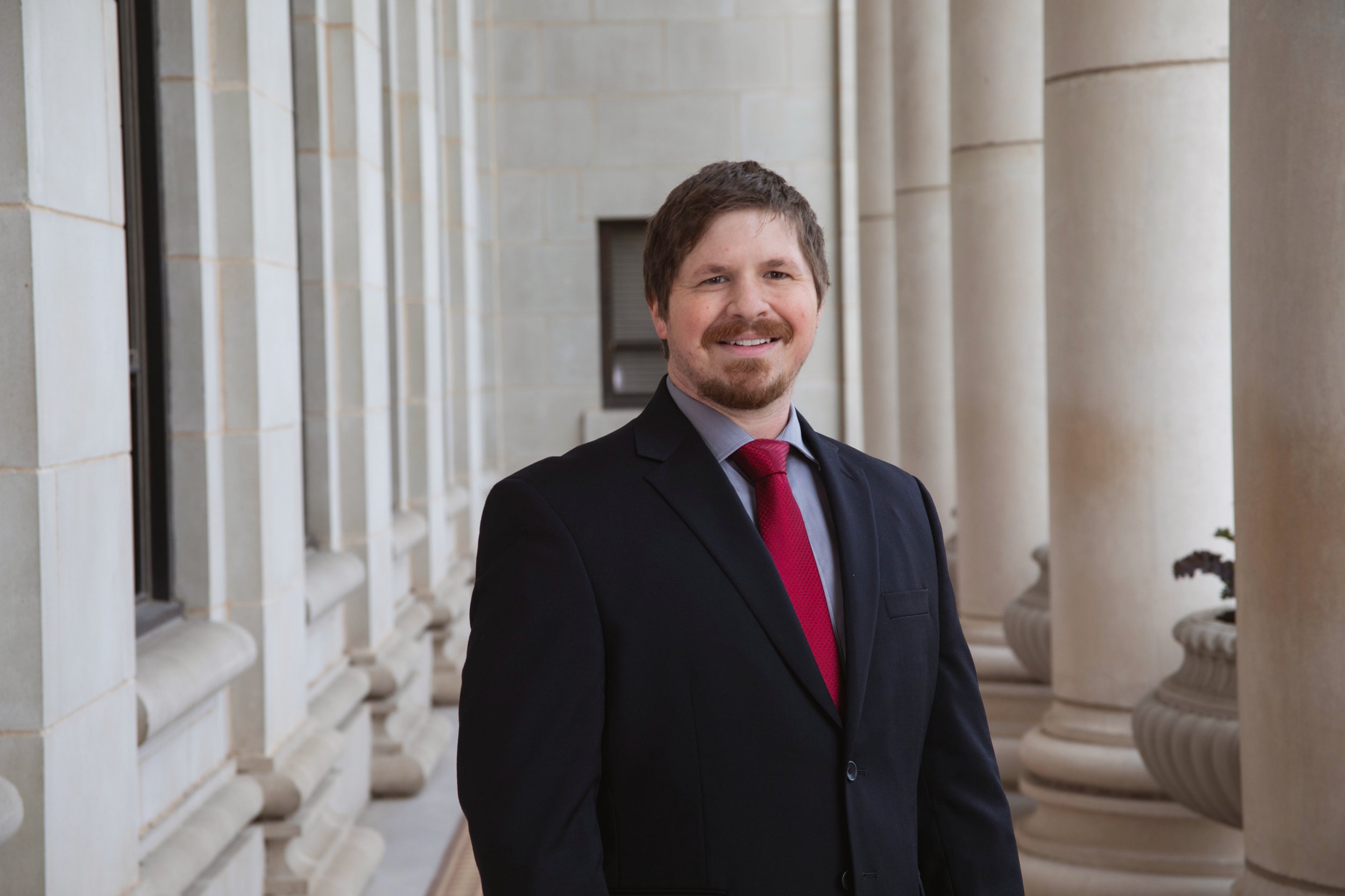 A man in a black suit with a red tie smiling, standing outdoors near a building with columns.