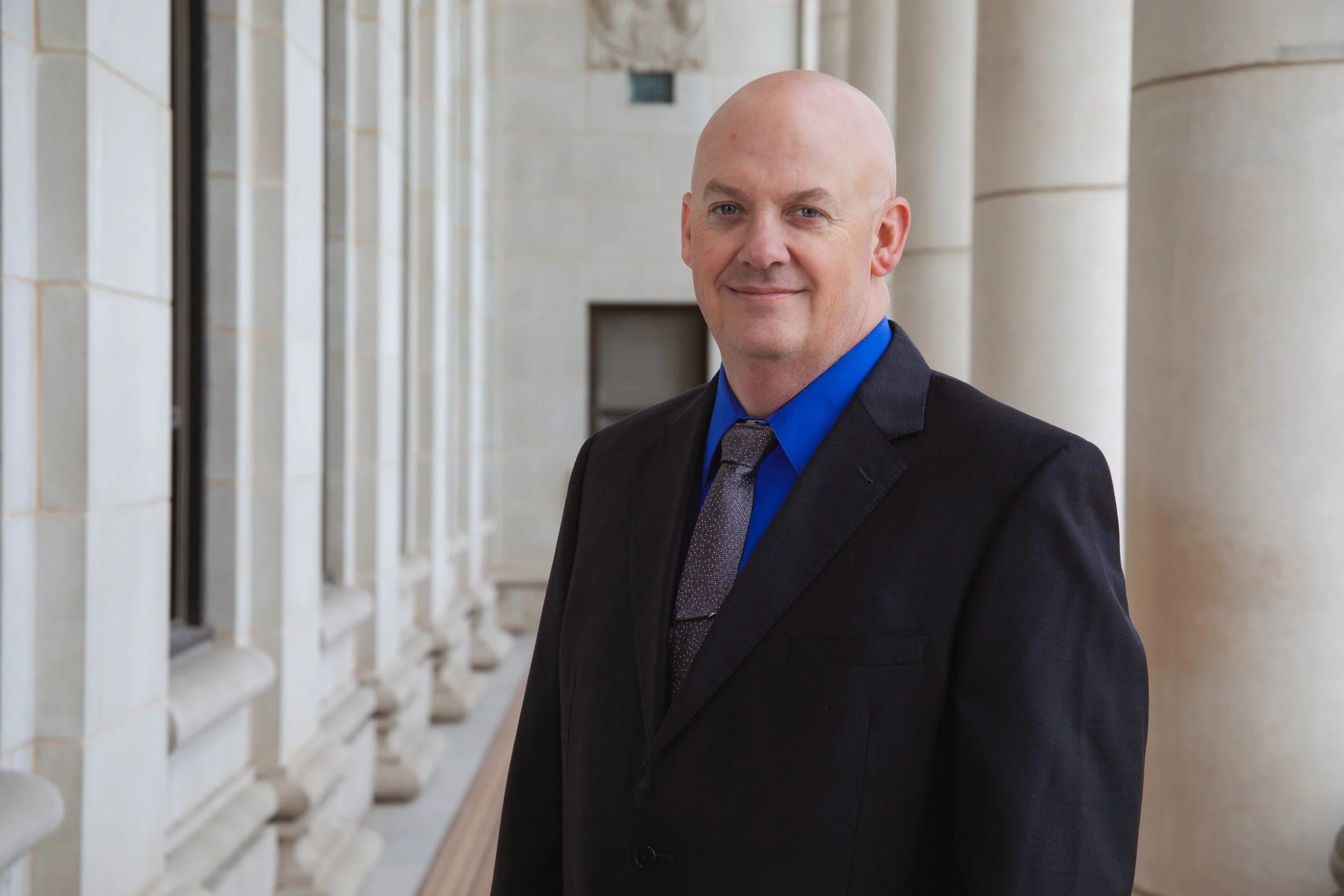 A bald man in a black suit, blue shirt, and patterned tie standing in a corridor with classical architecture, columns, and a sculpture on the wall behind him.