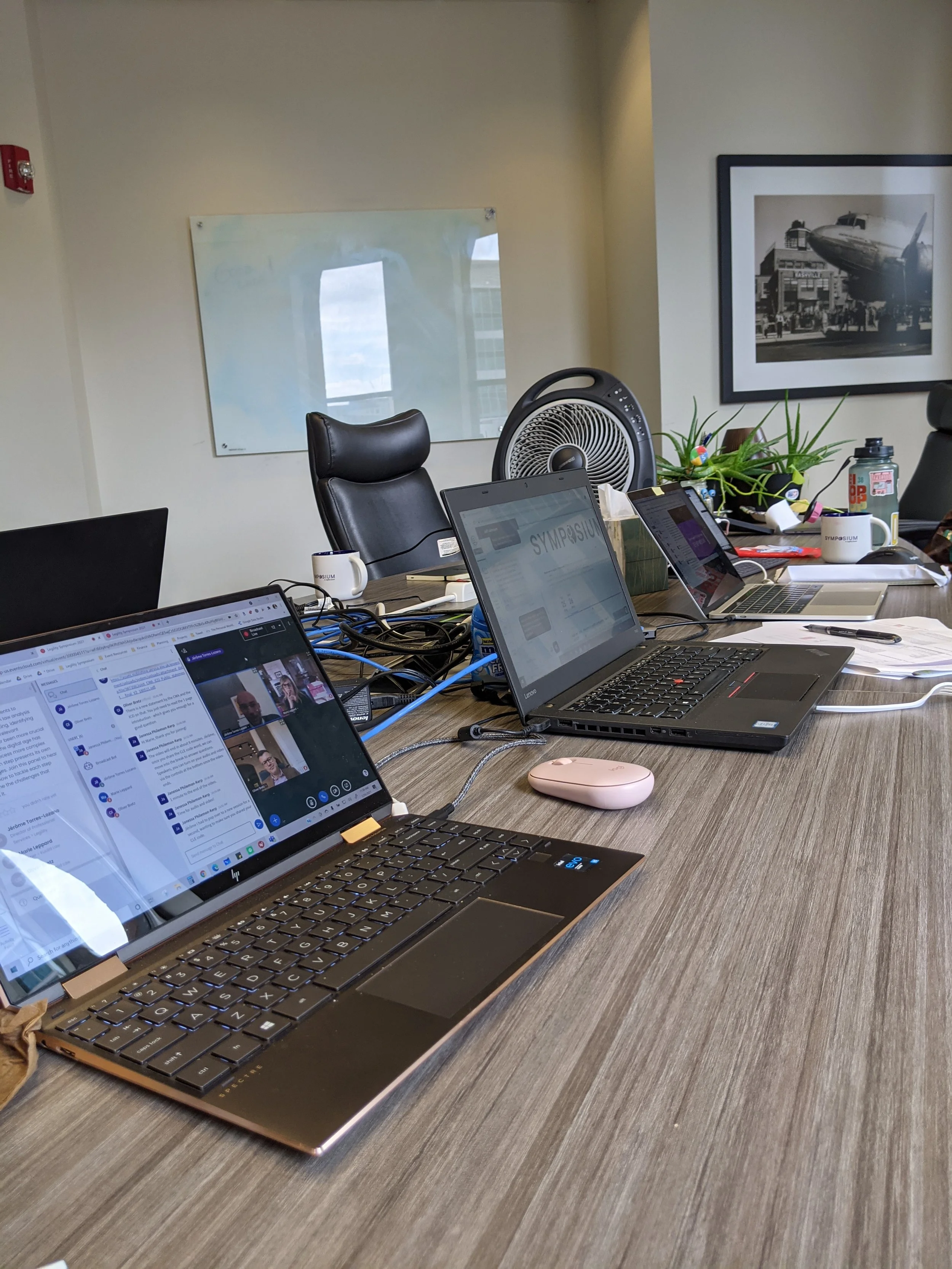 A laptop computer on a desk displaying a video call with two women, one Asian and one Caucasian, in a home setting. The desk has papers and a small container with a pair of glasses, and there is a background of houseplants and furniture.