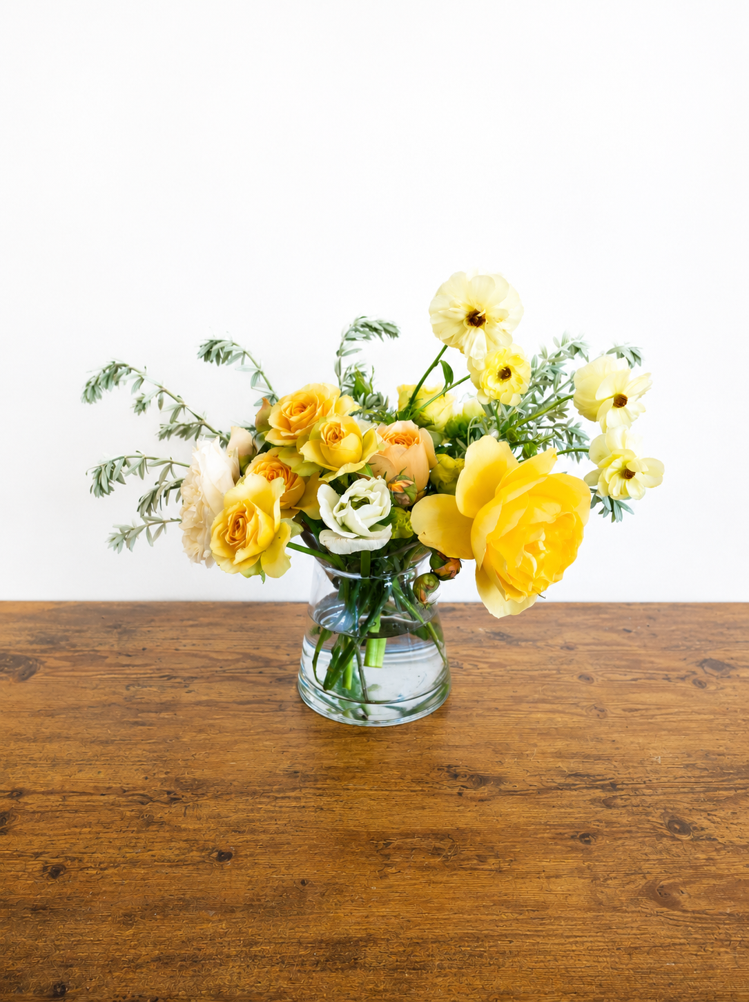 A glass vase with yellow, white, and cream-colored flowers on a wooden table against a plain white wall.