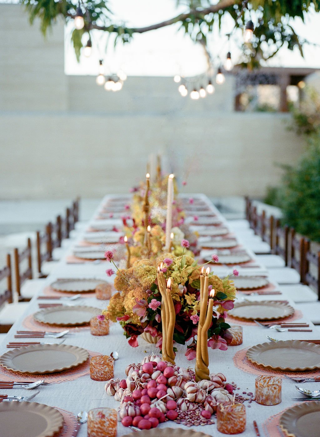 A long outdoor dining table decorated with pink, yellow, and purple flowers, candles, and gold accents under hanging string lights for an elevated dining experience.