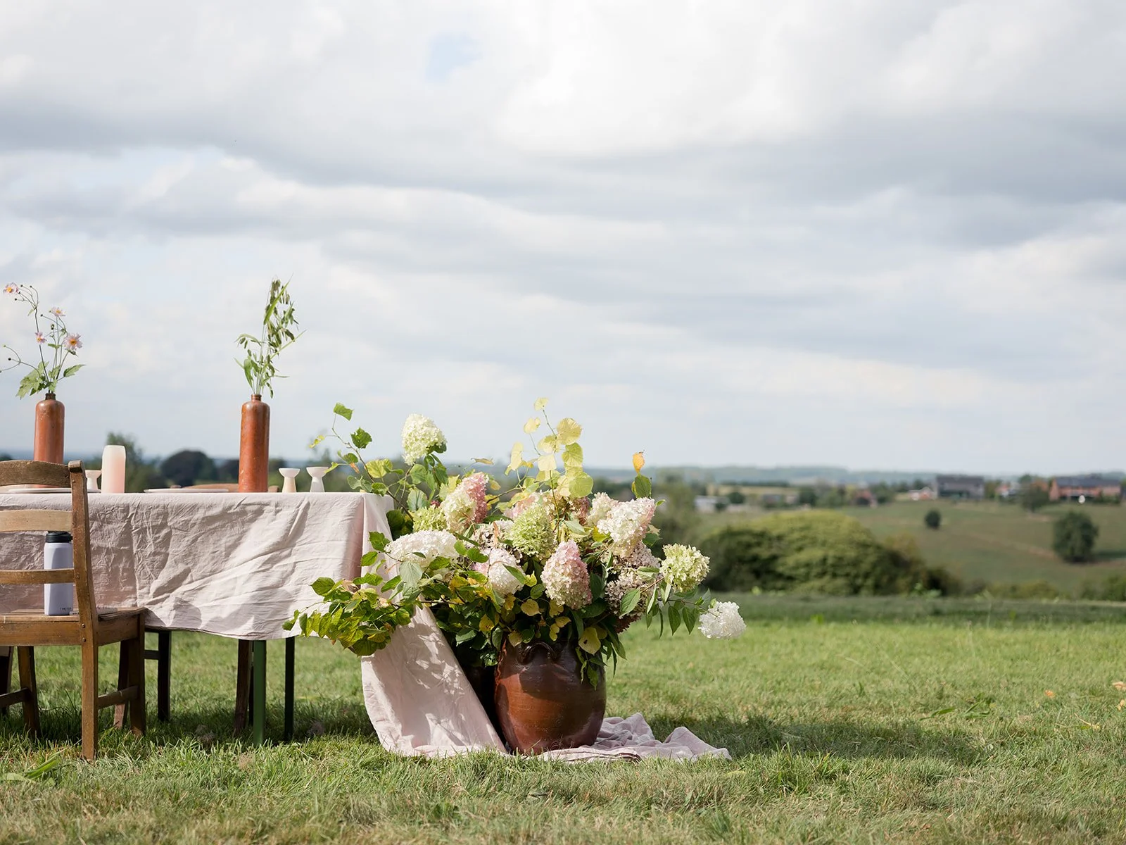 Outdoor dining setup with a table covered in a white tablecloth, decorated with vases of flowers, candles, and surrounded by a large flower arrangement on a grassy field with a scenic rural landscape under a cloudy sky.
