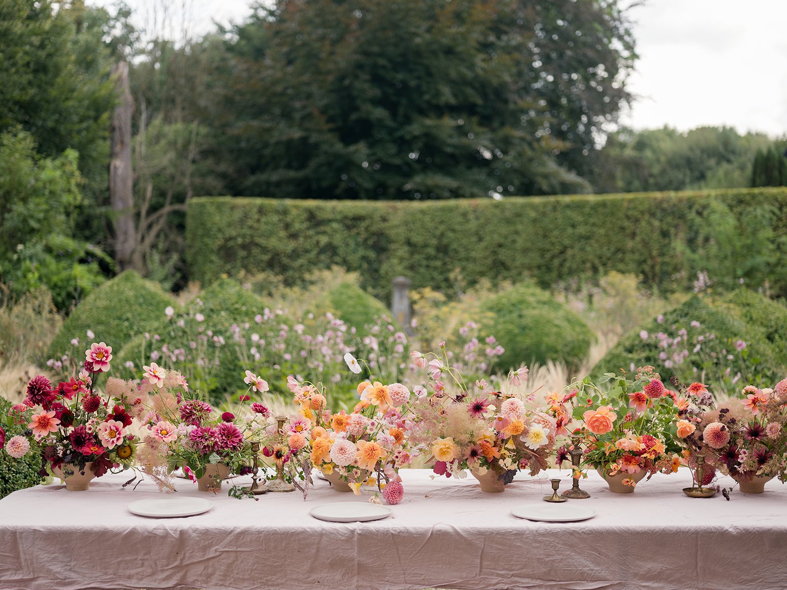 A long dining table draped with a pink tablecloth, decorated with multiple floral arrangements in vases, set outdoors with a lush green hedge and trees in the background.