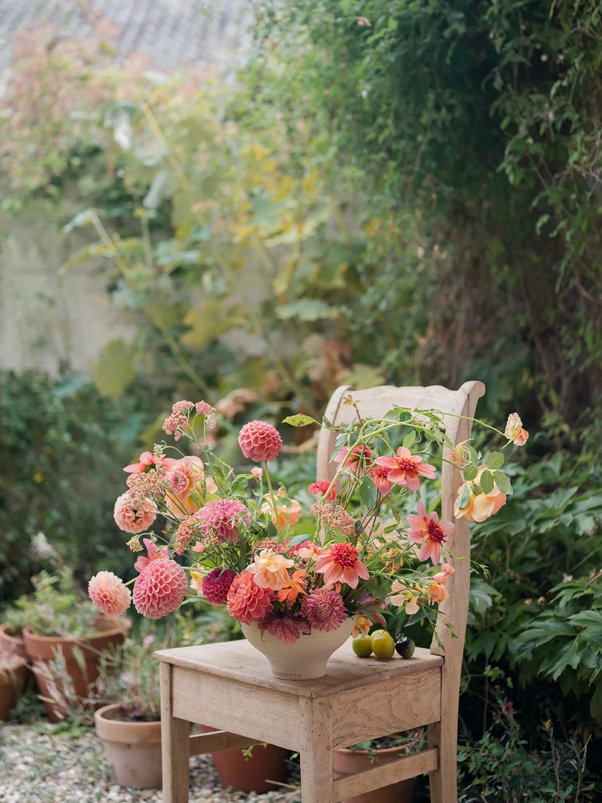 A wooden chair holding a large white vase filled with pink and peach flowers, with a few green plums on a small wooden table in front. The scene is set in a lush outdoor garden with greenery and potted plants.